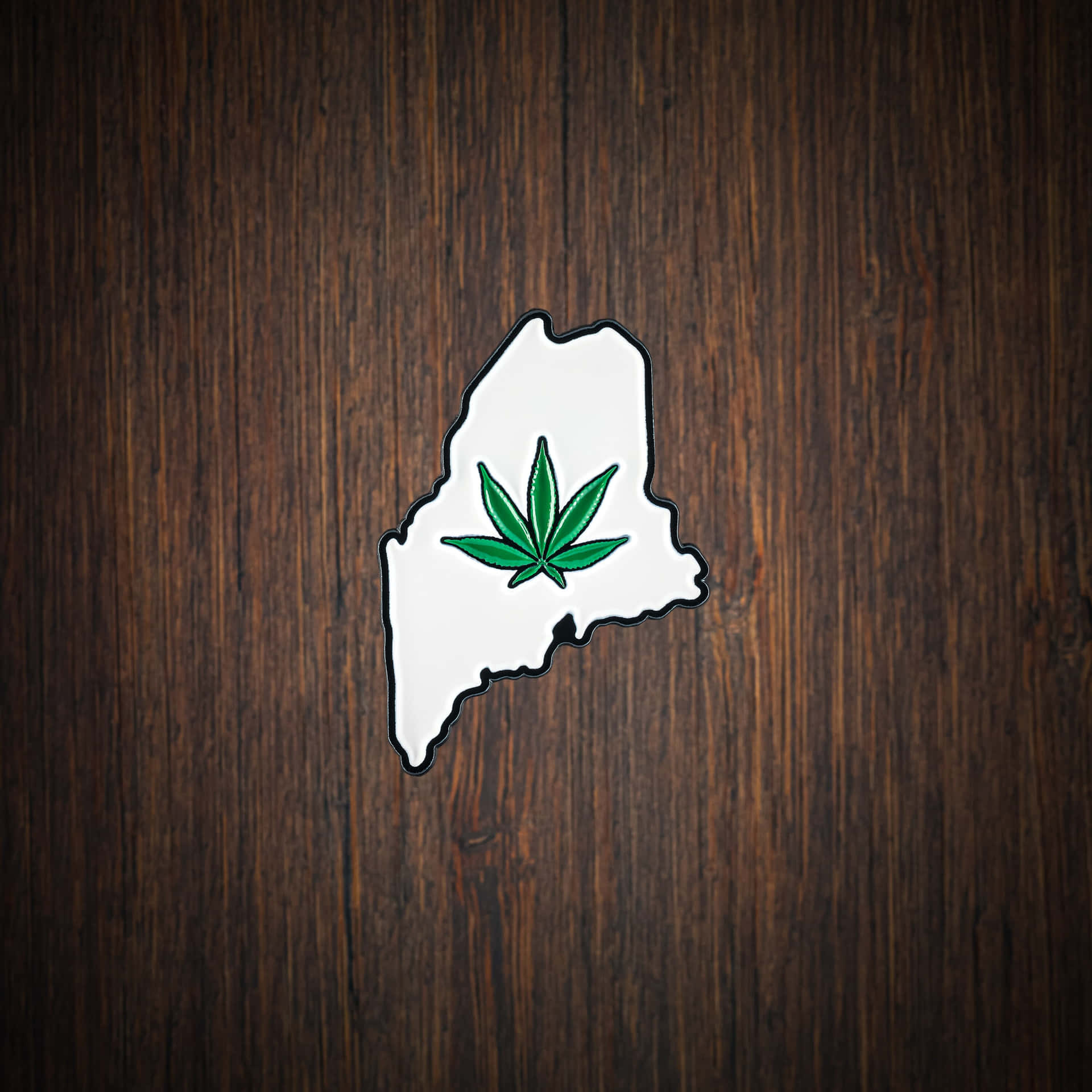  壁紙 — 一個白色琺瑯別針，形狀為麻薩諸塞州，中央有一個綠色大麻葉，置於深色木質背景上。