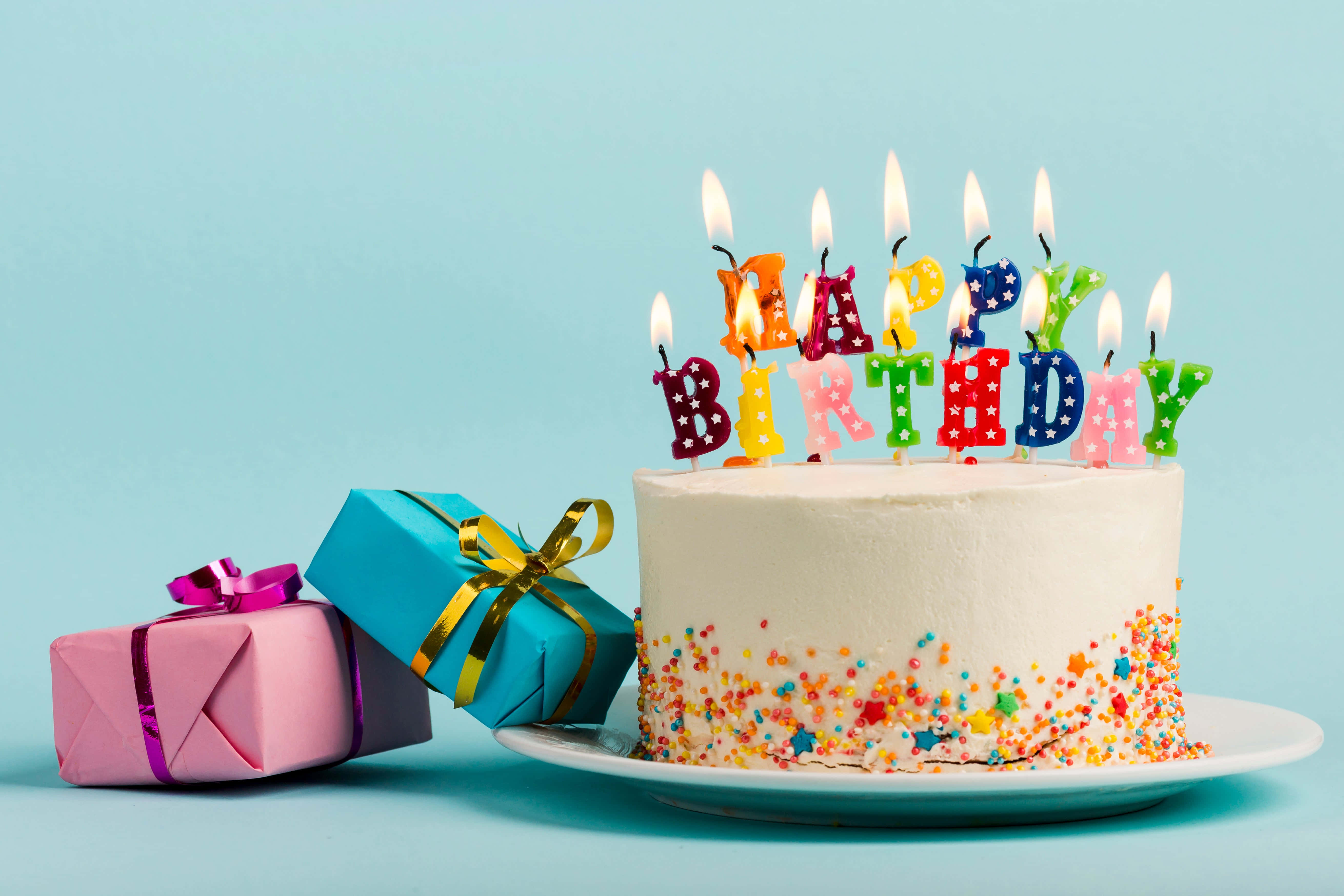 Birthday Cake 桌布 壁紙 — 一個白色生日蛋糕，上面有彩色字母蠟燭拼出 'HAPPY BIRTHDAY' 和星星糖粒，旁邊有兩個包裝好的禮物，放在淺藍色背景上。