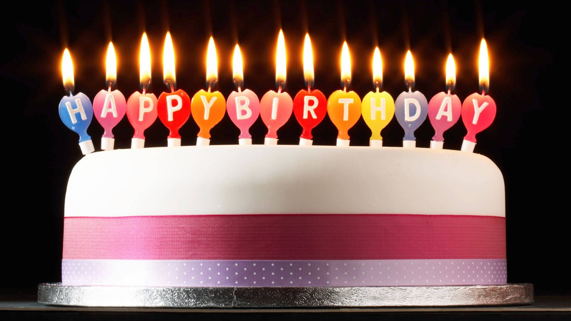 Birthday Cake 桌布 壁紙 — 一個白色生日蛋糕，上面有彩色心形蠟燭拼出「生日快樂」，背景為黑色。