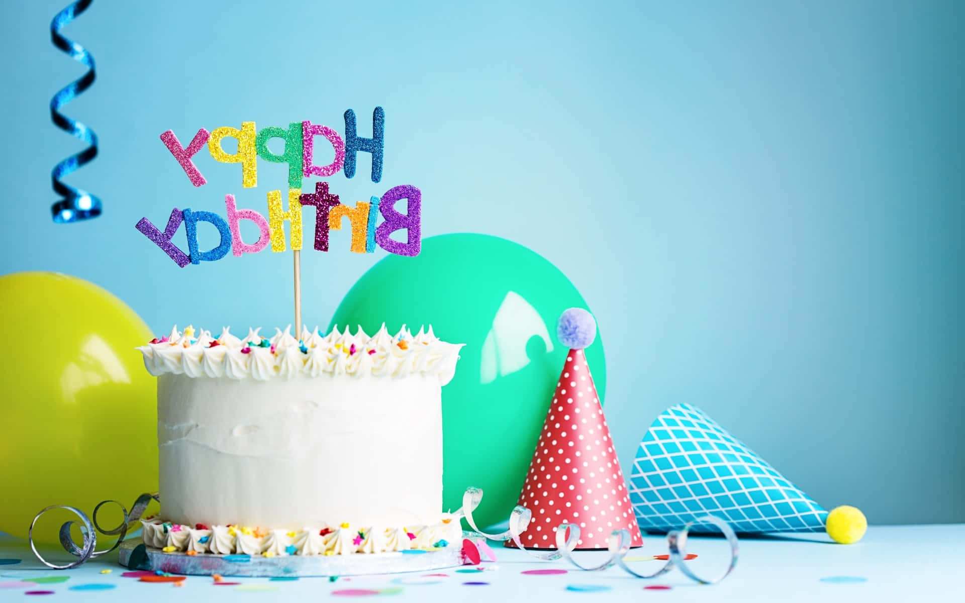 Birthday Cake 桌布 壁紙 — 一個白色生日蛋糕，上面有彩色糖粒和閃亮的「Happy Birthday」裝飾，周圍環繞著氣球和派對帽。