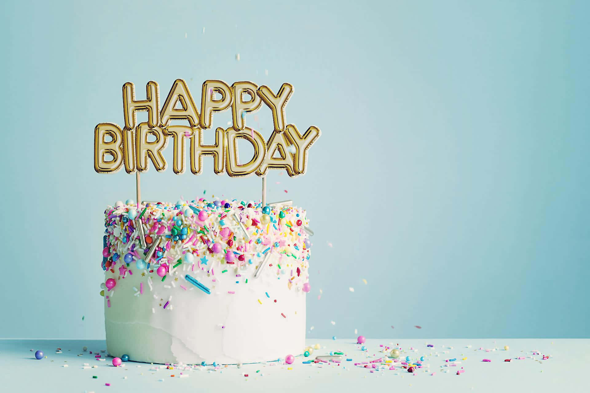 蛋糕 桌布 壁紙 — 一個白色生日蛋糕，上面有金色「HAPPY BIRTHDAY」字母和彩色糖粒，襯托柔軟的藍色背景。