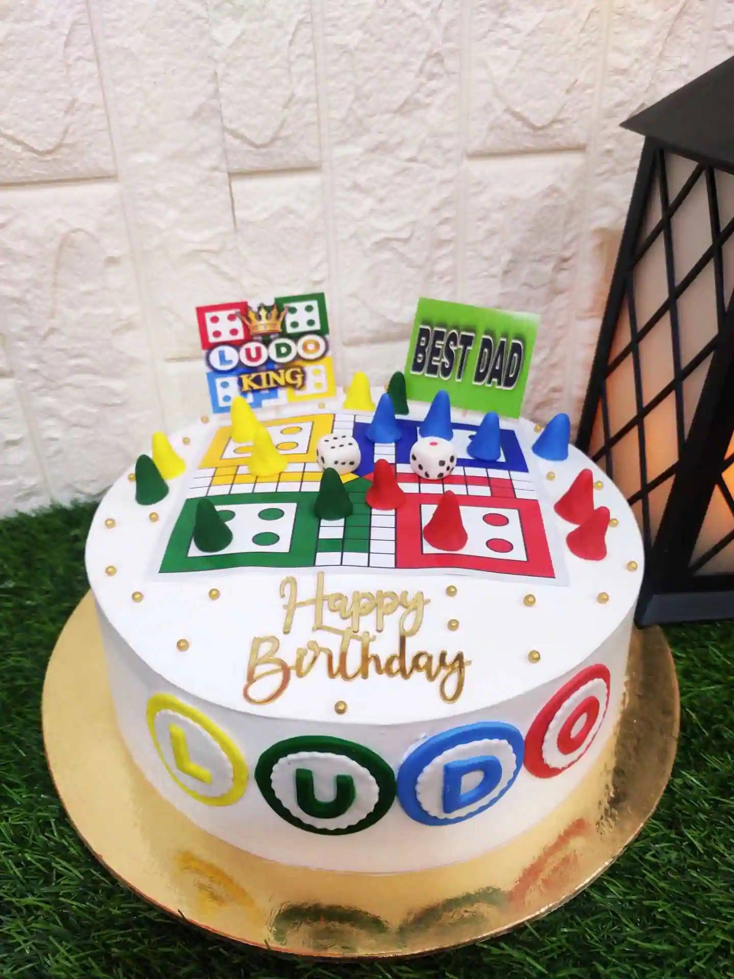 桌布 壁紙 — 一個白色生日蛋糕，裝飾著色彩繽紛的Ludo棋盤遊戲主題，並有「最佳爸爸」裝飾，放在綠色草地上。