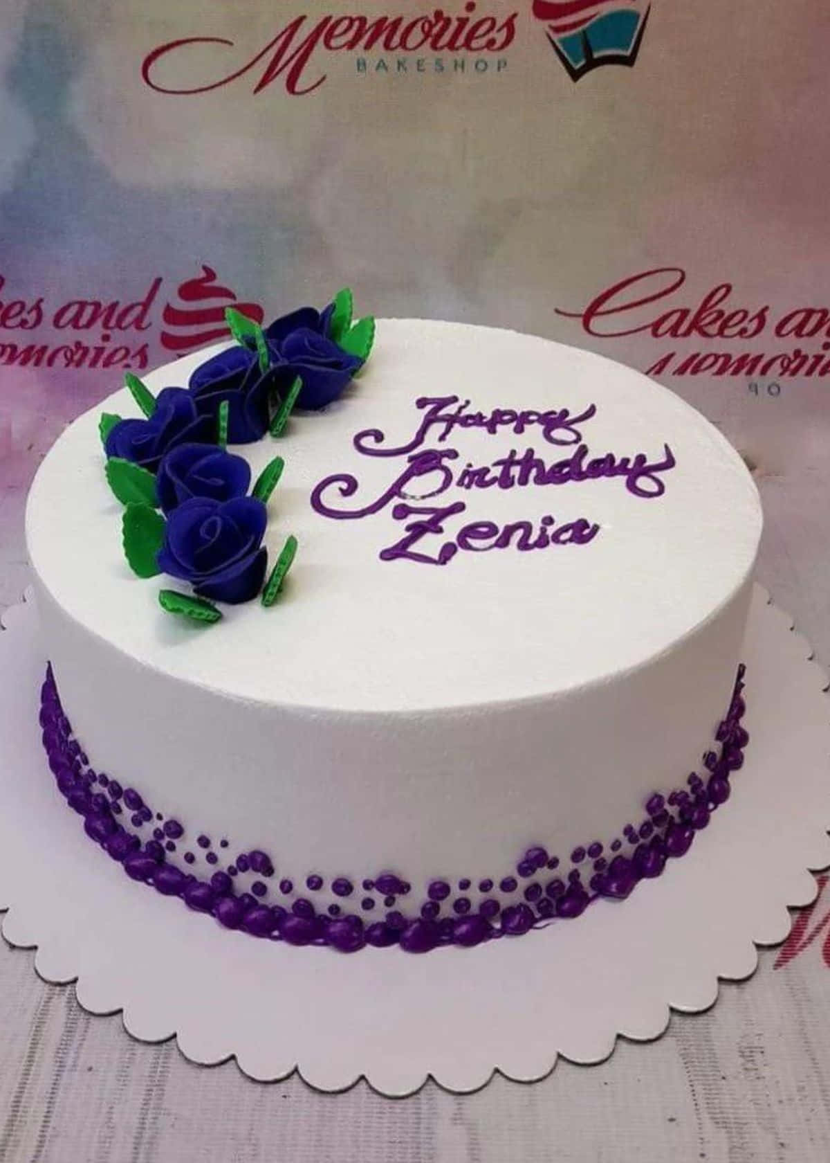 Floral Cake 桌布 壁紙 — 一個白色生日蛋糕，飾有紫色玫瑰和紫色字體寫著「Happy Birthday Zenia」，放在一個扇貝邊的托盤上。