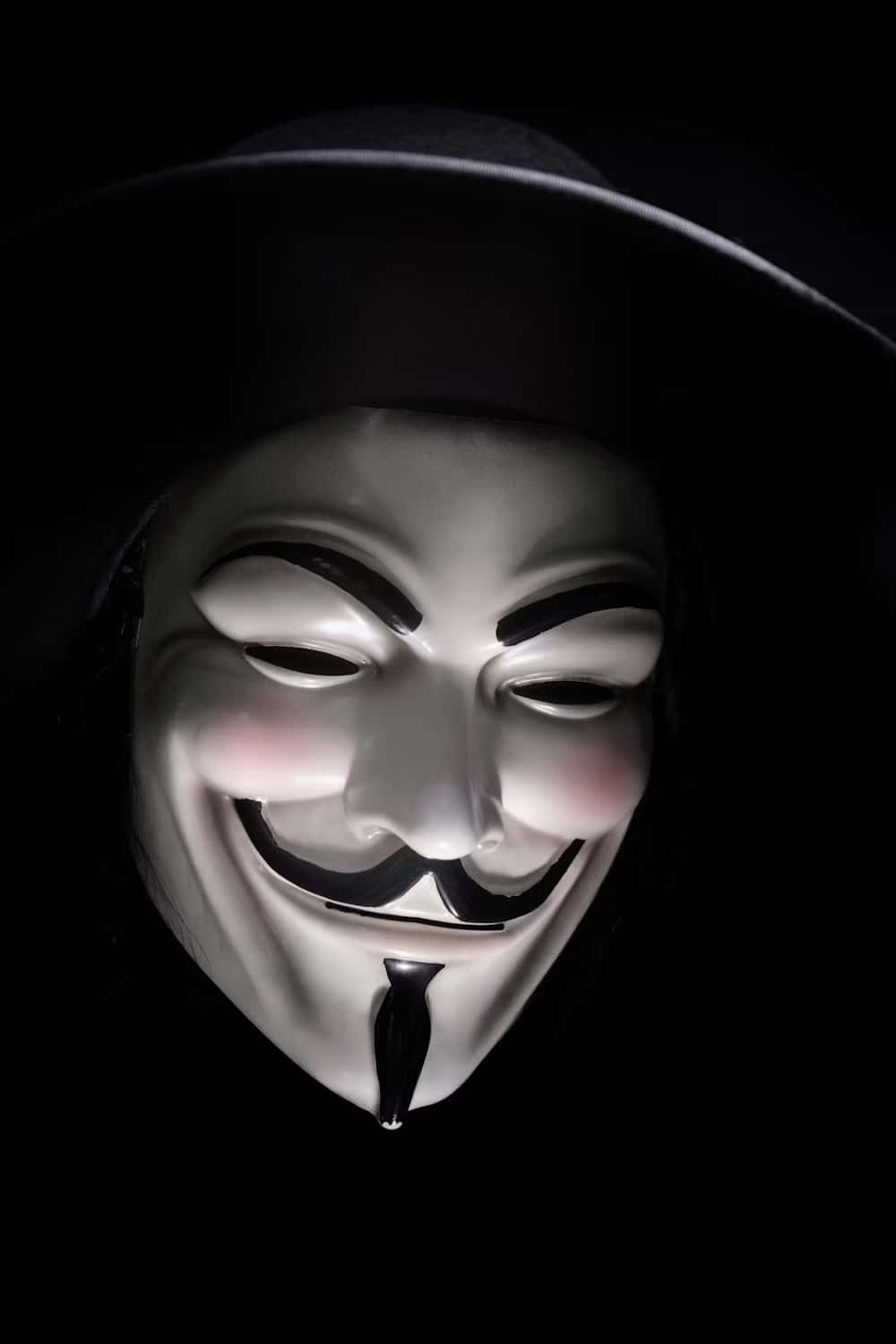  壁紙 — 一個白色的 Guy Fawkes 面具，帶著一個大大的笑容和黑色帽子，在漆黑的背景前被照亮。