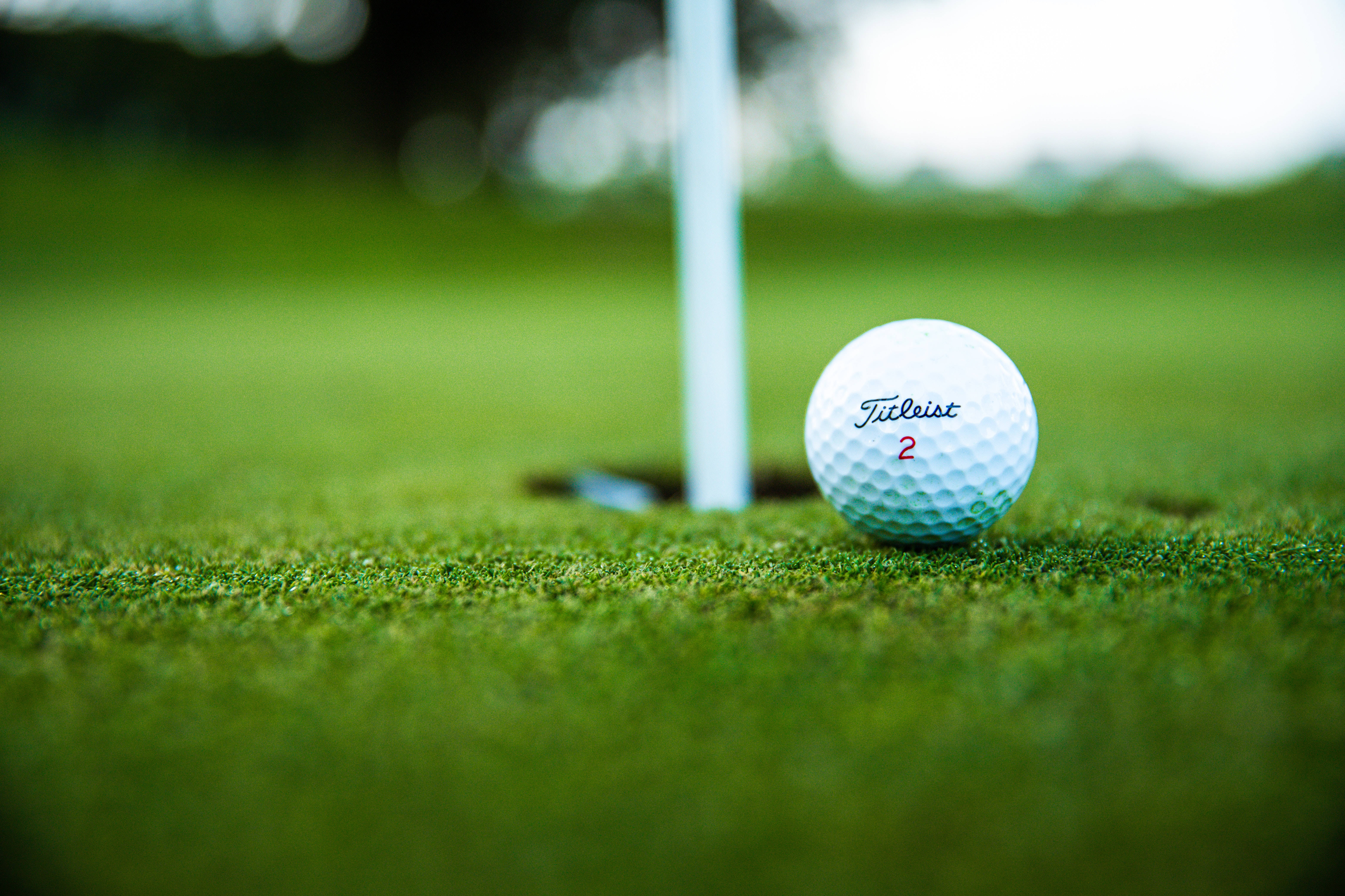 4K Golf 桌布 壁紙 — 一個白色的Titleist高爾夫球放在鮮綠色的草地上，旁邊是一個帶有白色旗桿的高爾夫球洞。