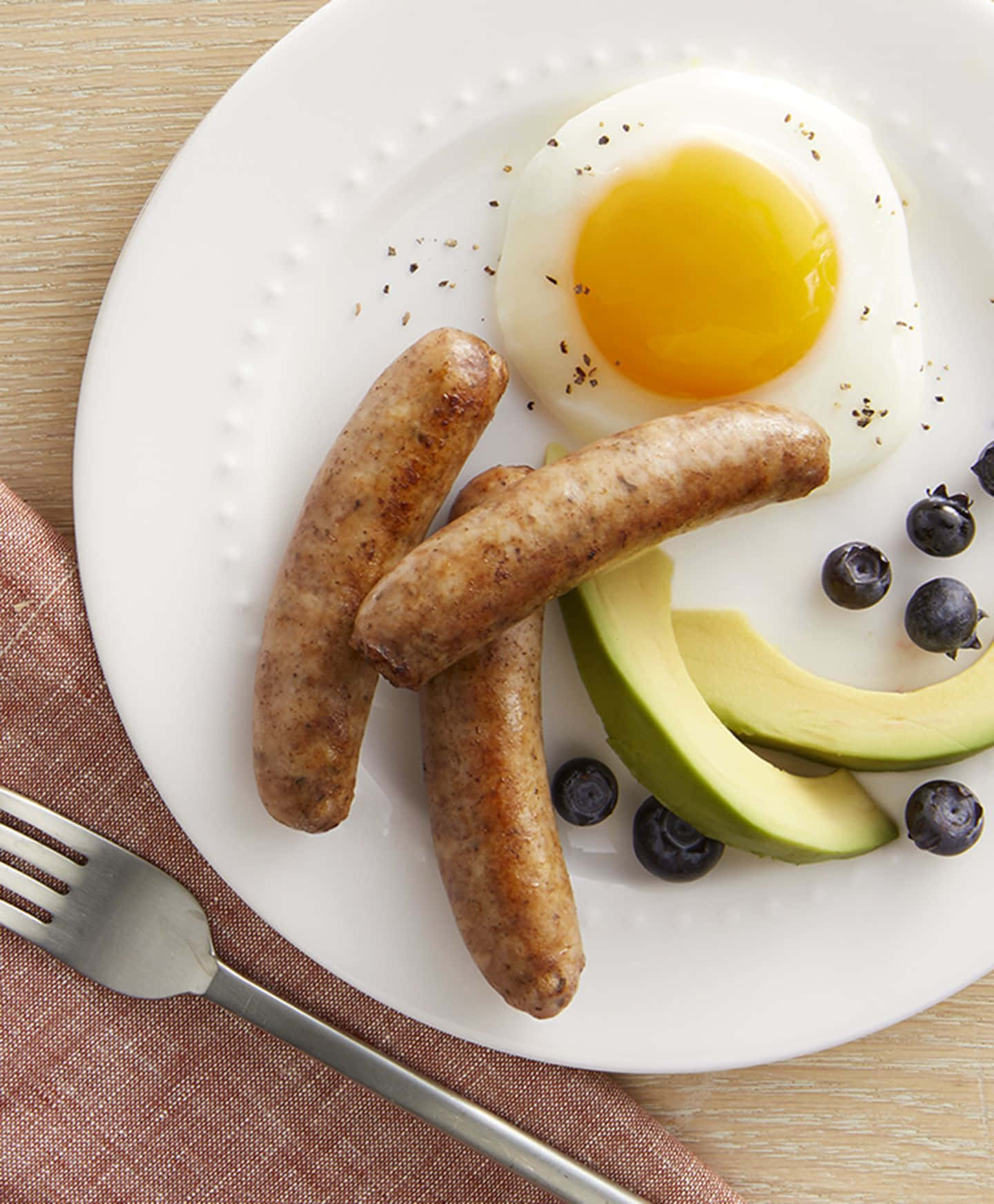 Sausage 桌布 壁紙 — 一個白色盤子上放著三條煎香腸、一顆太陽蛋、酪梨片和藍莓，放在木桌上，旁邊有叉子。