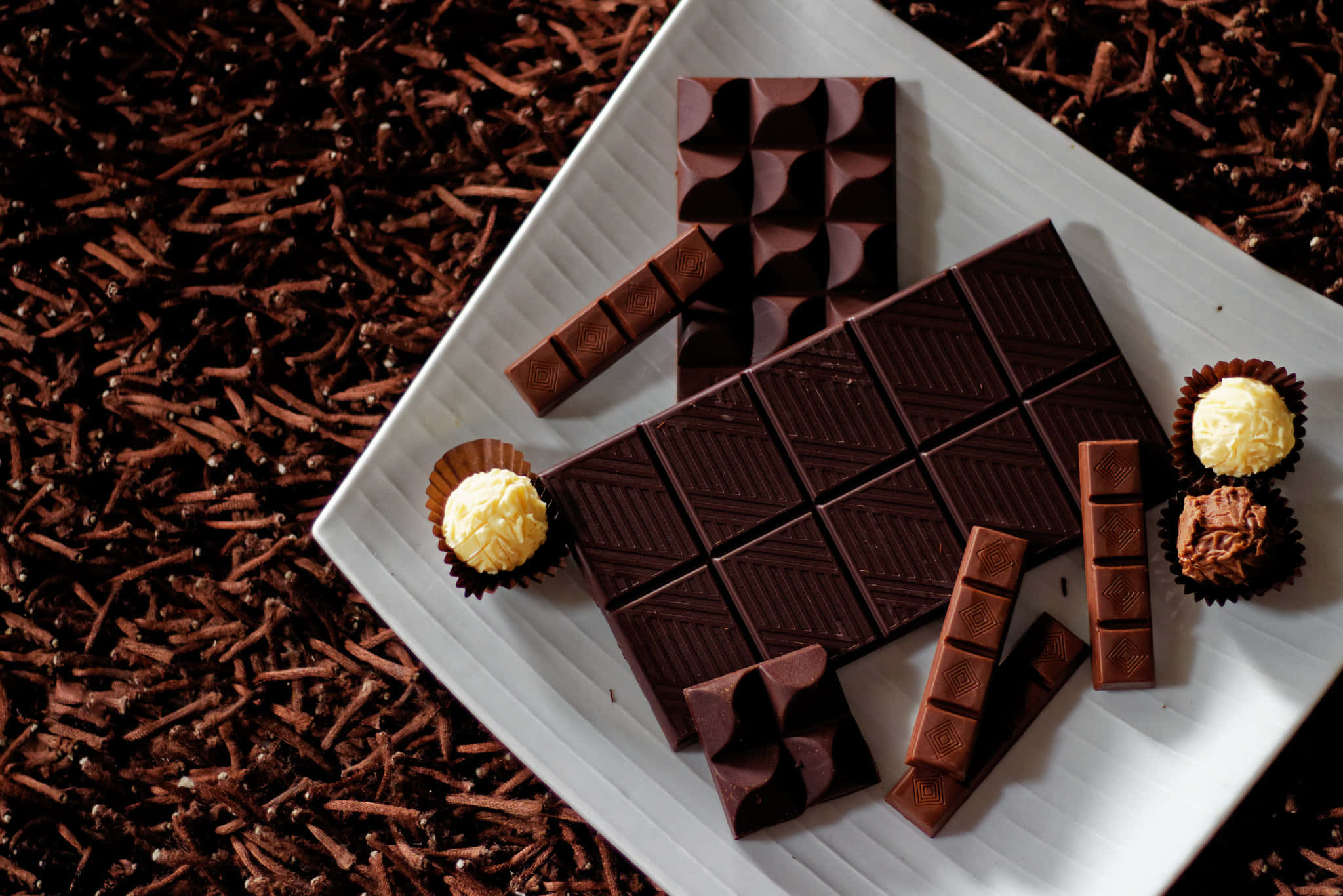 Dark Chocolate 桌布 壁紙 — 一個白色盤子上放著各式各樣的黑巧克力條和松露巧克力，周圍是可可碎粒，背景是豐富的棕色。