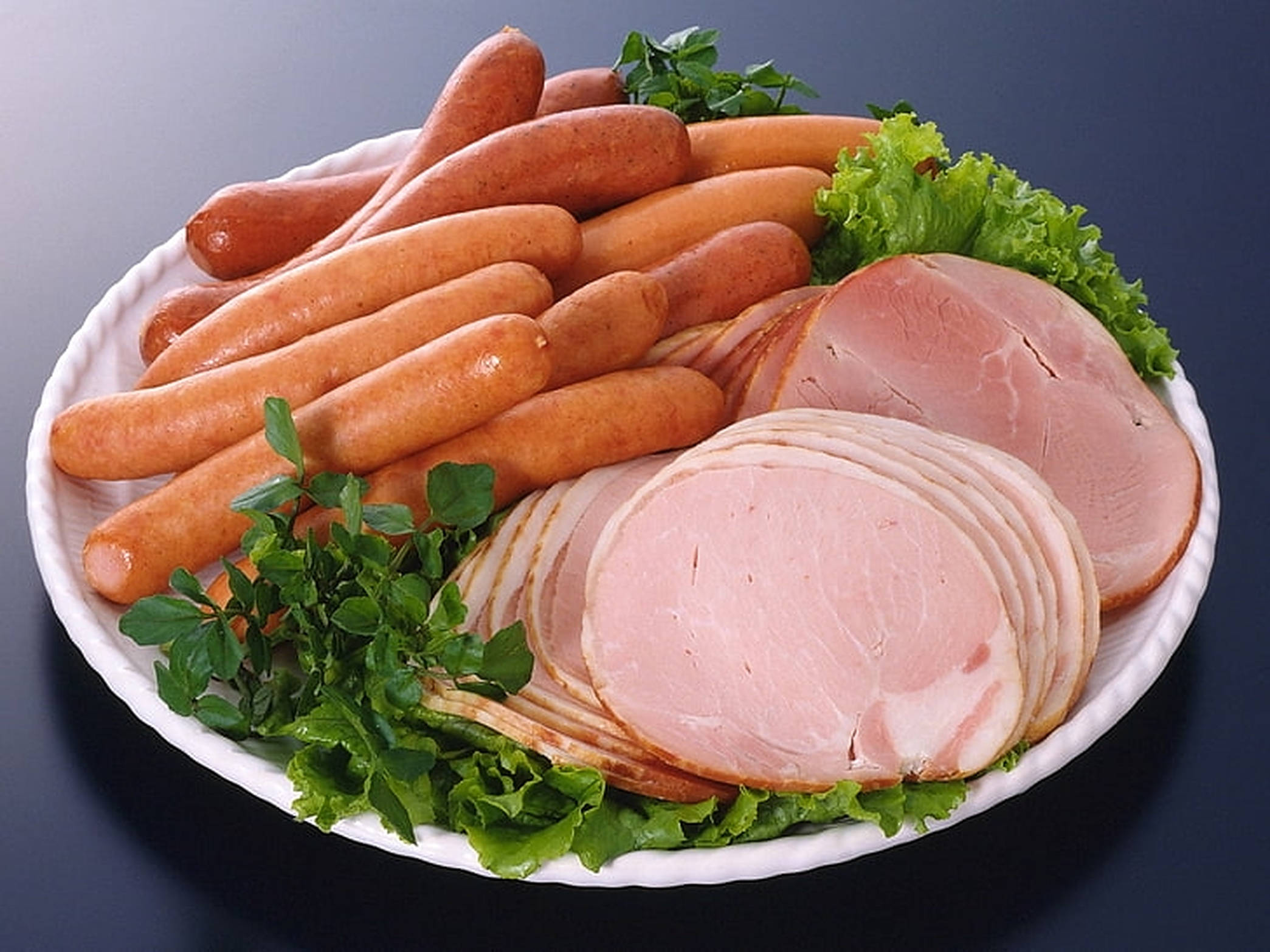 Ham 桌布 壁紙 — 一個白色盤子上放著金黃色的香腸和切片火腿，配上新鮮的綠色生菜和歐芹，背景為深色。