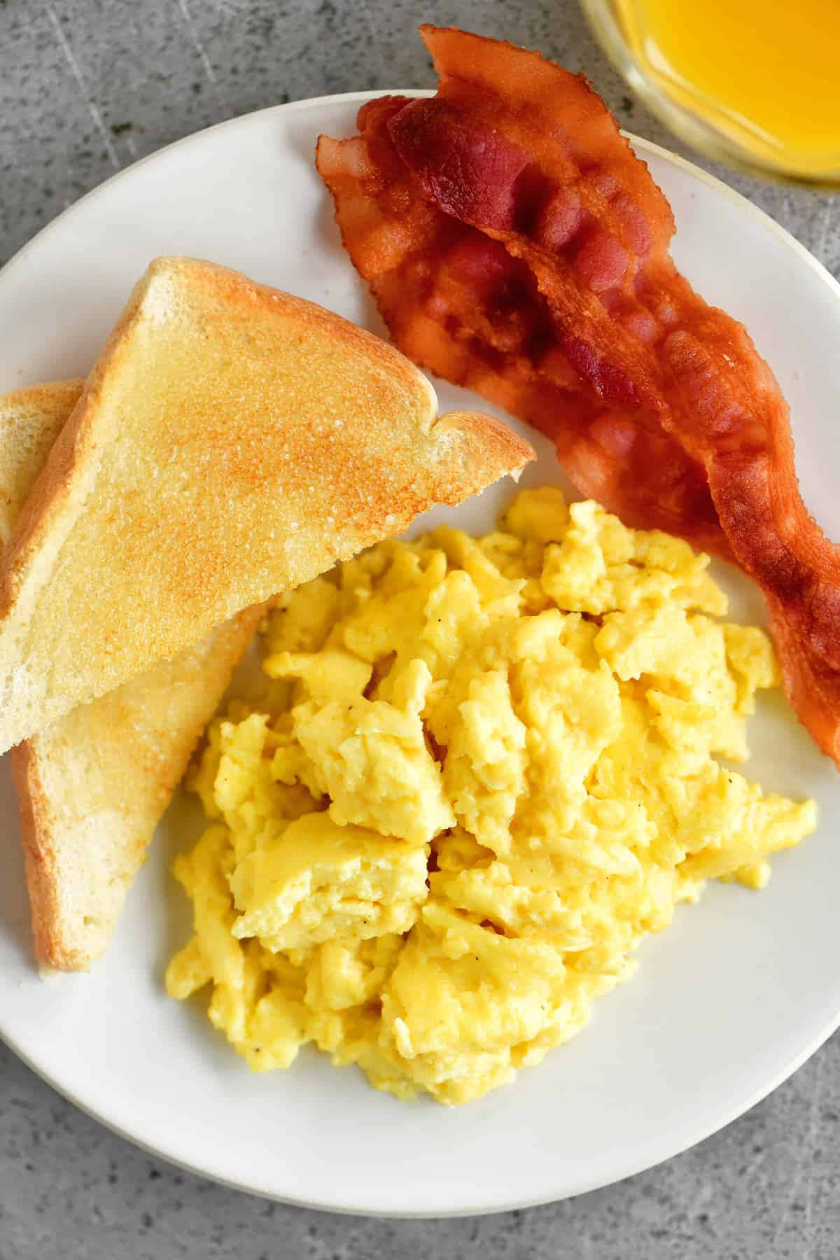 Scrambled Egg 桌布 壁紙 — 一個白色盤子的特寫，上面堆滿了鬆軟的炒蛋、酥脆的培根和金黃色的吐司，旁邊有一杯柳橙汁。