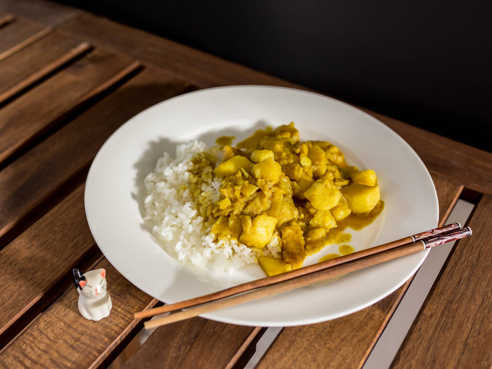 Food Photography 桌布 壁紙 — 一個白色盤子盛裝著冒煙的黃咖哩、馬鈴薯和白飯，放在木桌上，旁邊有筷子和一個小貓公仔。