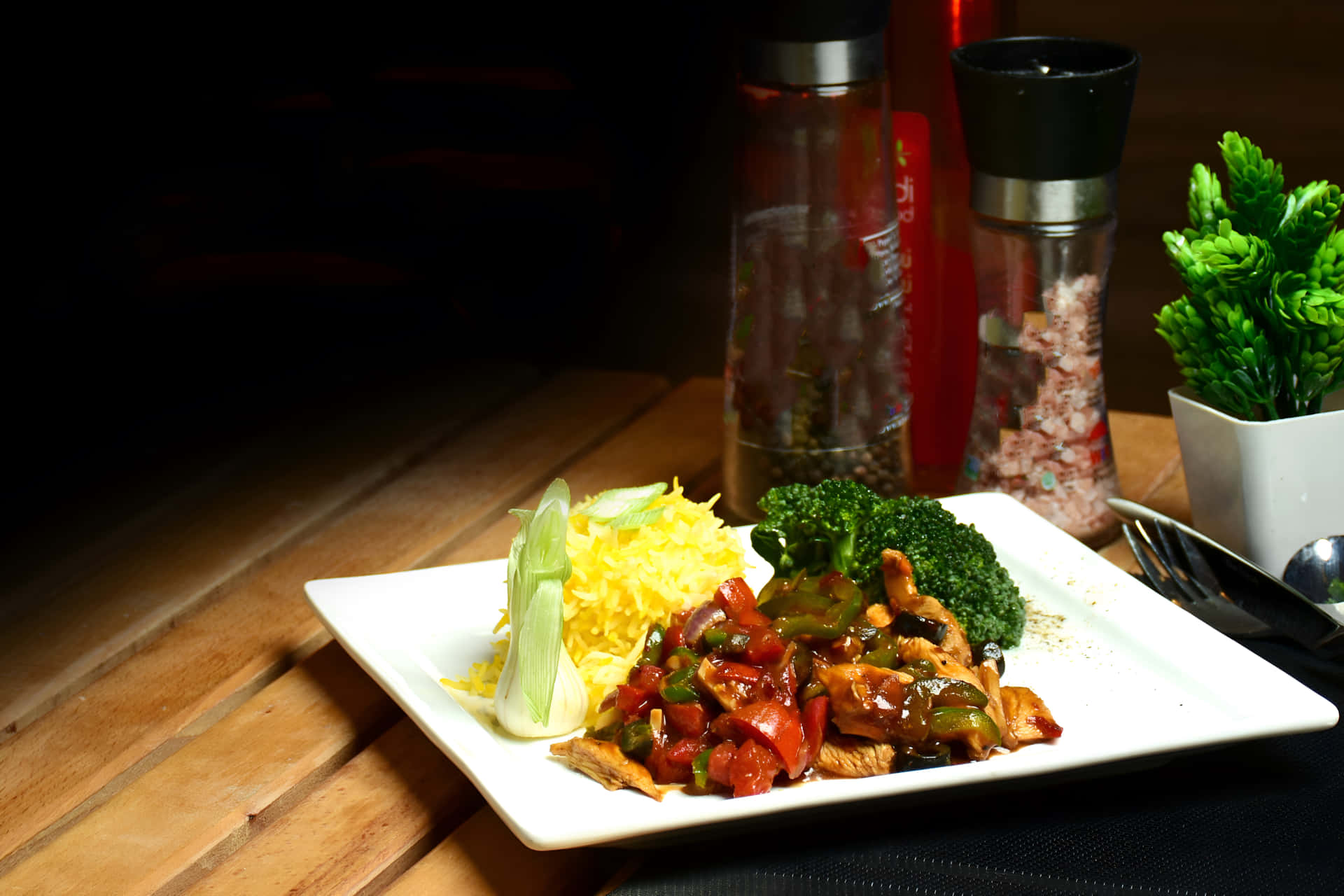 Chicken Chilli Dry with rice on a plate 壁紙 — 一個白色盤子盛裝著炒雞肉花椰菜和黃色米飯，放在木桌上，旁邊有鹽和胡椒罐以及一盆綠色植物。