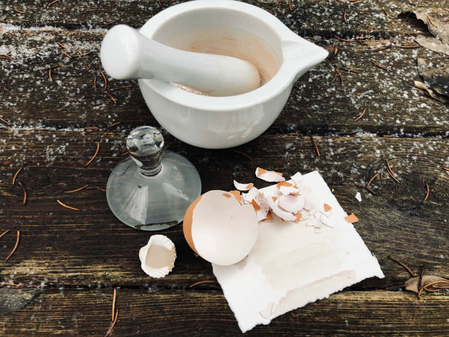 Eggshell 桌布 壁紙 — 一個白色研缽和研杵，內有壓碎的粉末，旁邊有破碎的蛋殼和一個玻璃杯，放在積雪的木質表面上。