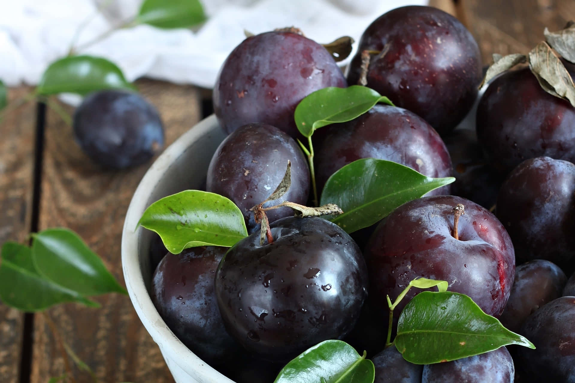 Purple Plums 桌布 壁紙 — 一個白色碗，裝滿了光澤的深紫色李子和新鮮的綠葉，放在質樸的木桌上。
