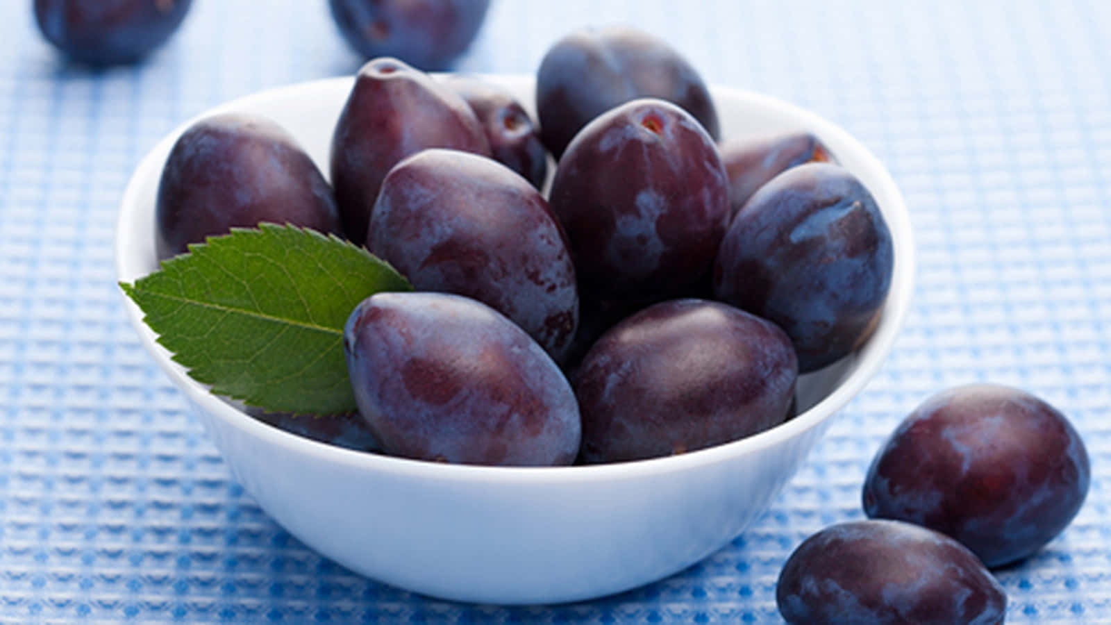 Fruits 桌布 壁紙 — 一個白色碗，裝滿飽滿的深紫色李子和新鮮的綠葉，放在藍色格子布上。
