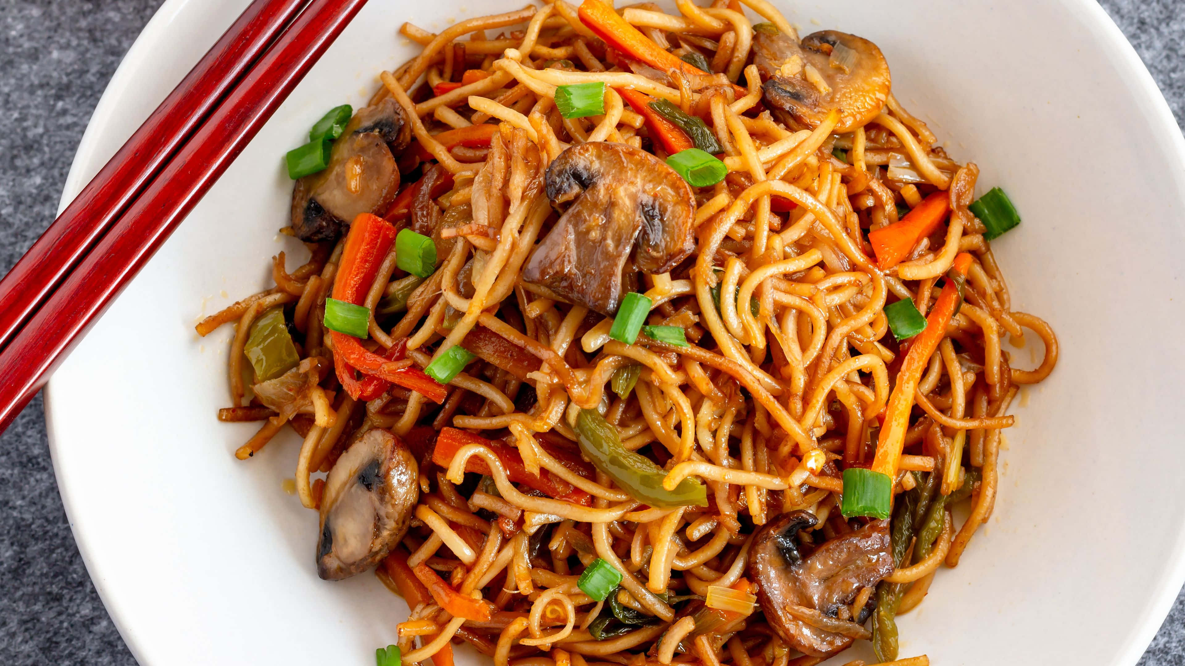 Noodles 桌布 壁紙 — 一個白色碗中的炒麵，搭配蘑菇、紅蘿蔔和青蔥，並有紅色筷子，特寫鏡頭。