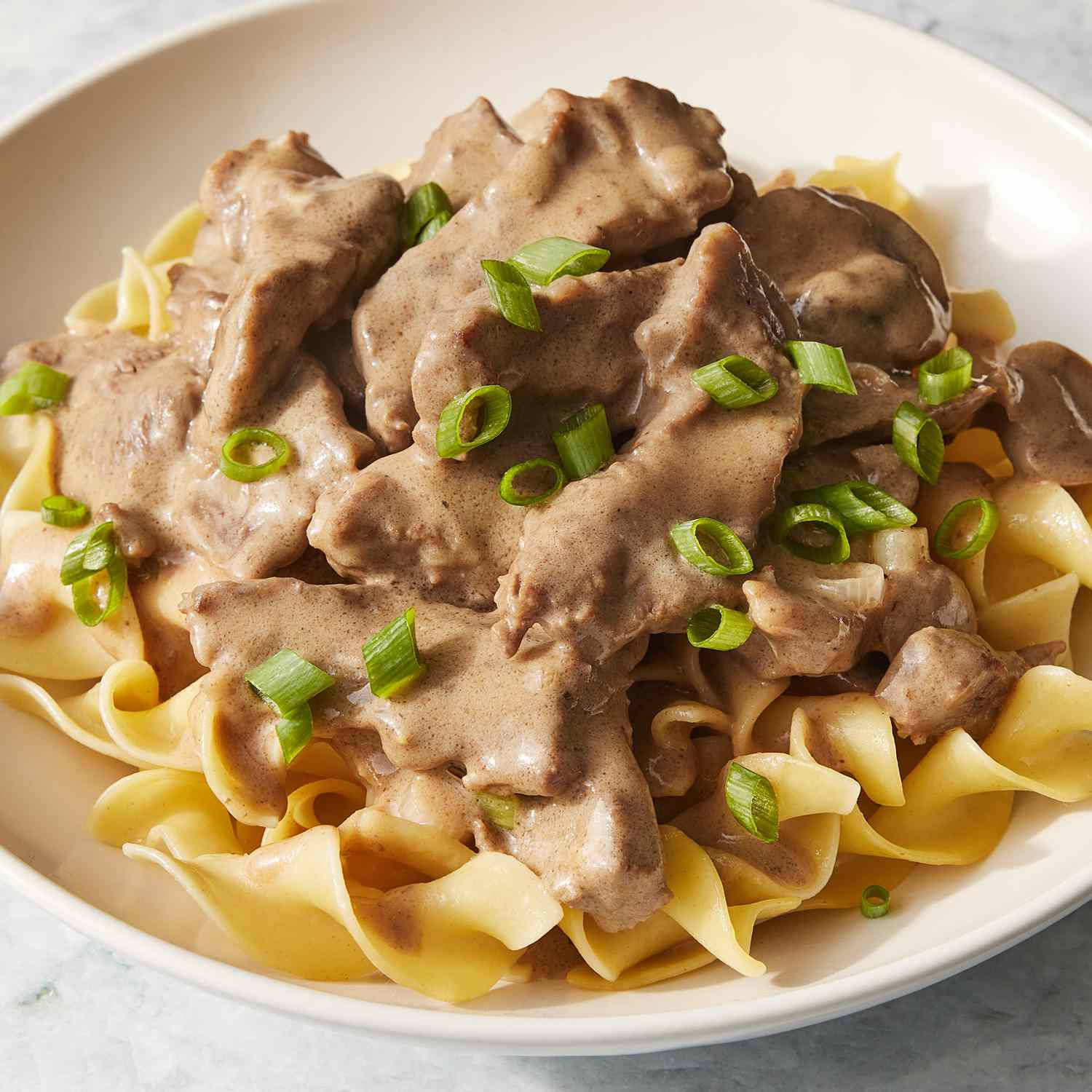 Food Photography 桌布 壁紙 — 一個白色碗中盛載著奶油牛肉斯特羅加諾夫，搭配切片蘑菇和切碎青蔥，放在寬蛋麵上。