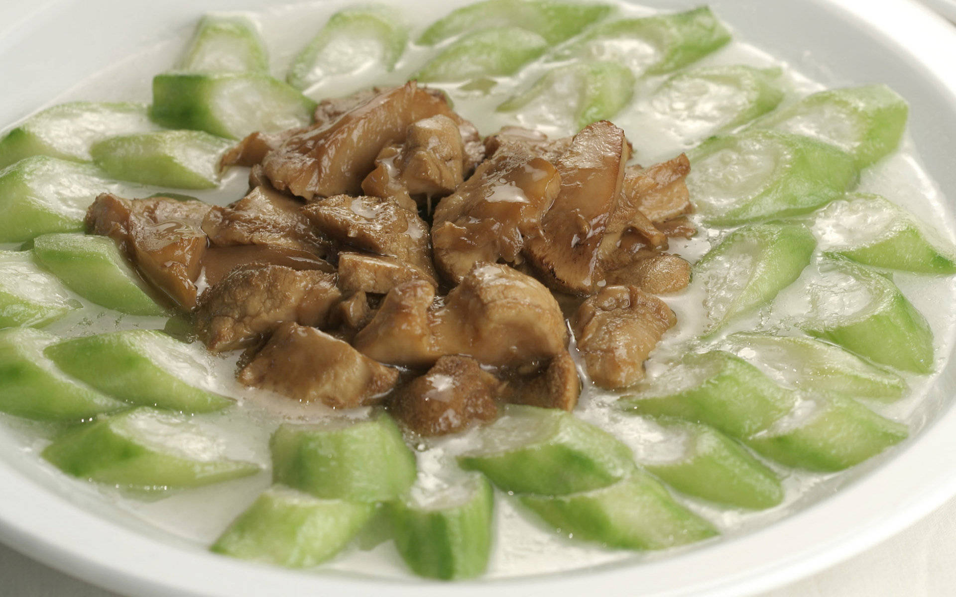 4K 食物 桌布 壁紙 — 一個白色碗中盛載著嫩滑的棕色肉塊，浸在奶油醬汁中，周圍環繞著鮮綠色的黃瓜片。