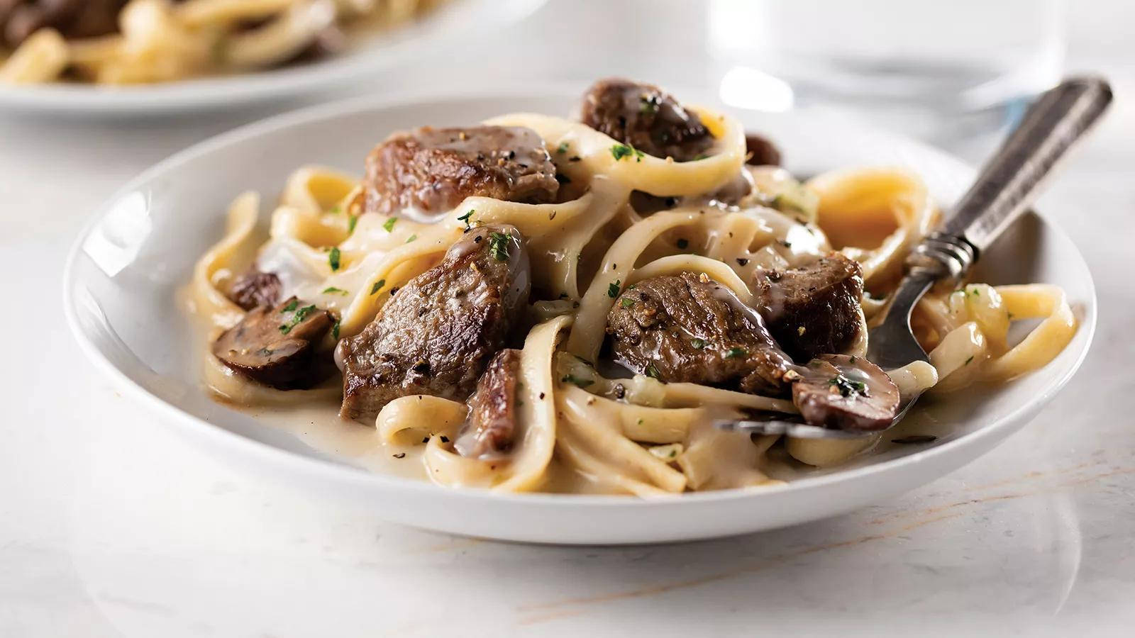 Pasta 桌布 壁紙 — 一個白色碗的特寫，裡面裝滿了奶油牛肉斯特羅加諾夫，放在寬麵條上，並用新鮮歐芹裝飾。