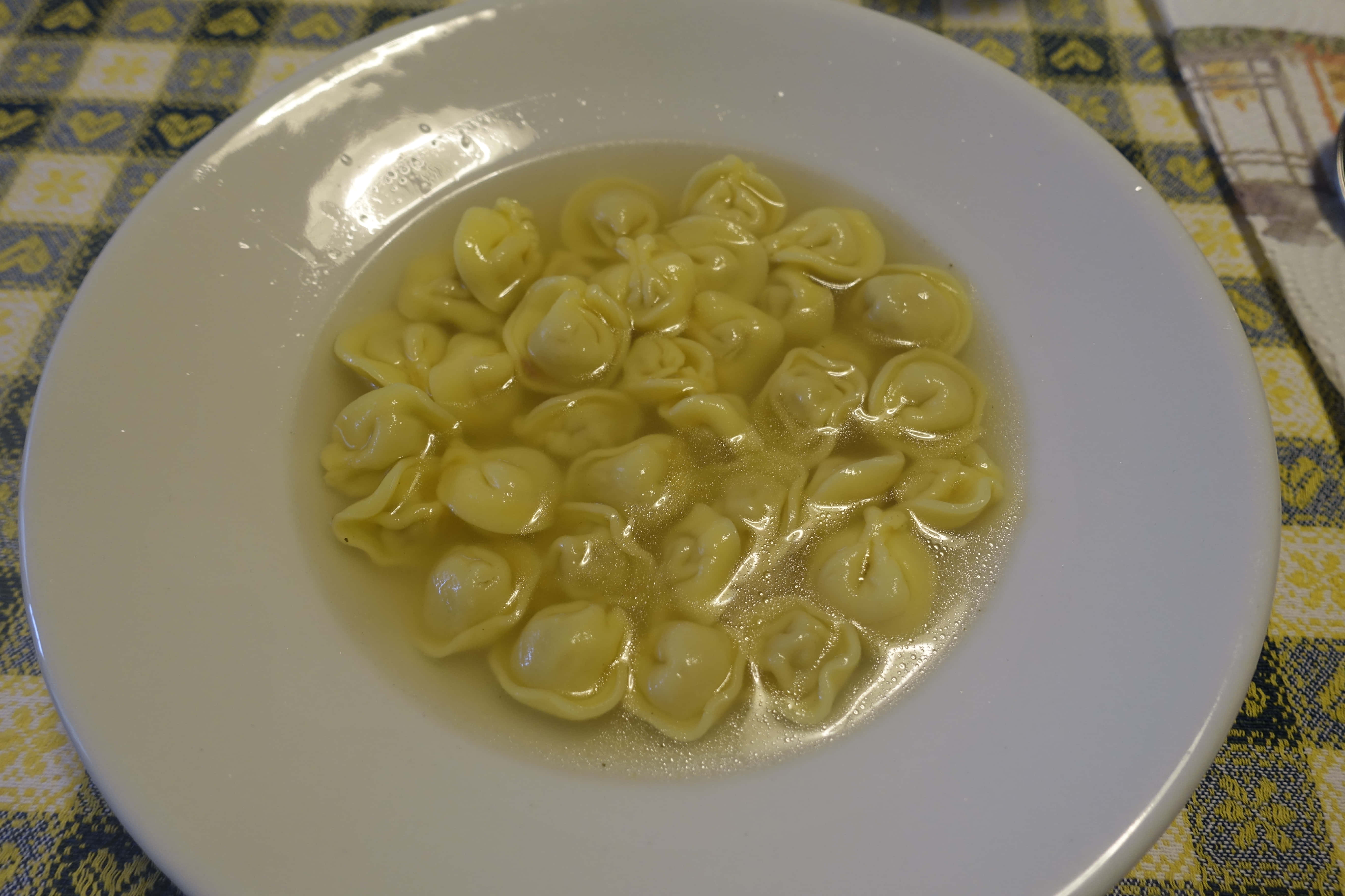 Pasta 桌布 壁紙 — 一個白色碗盛滿飽滿的黃色義大利餃子在清湯中，放在黃藍圖案桌布上。