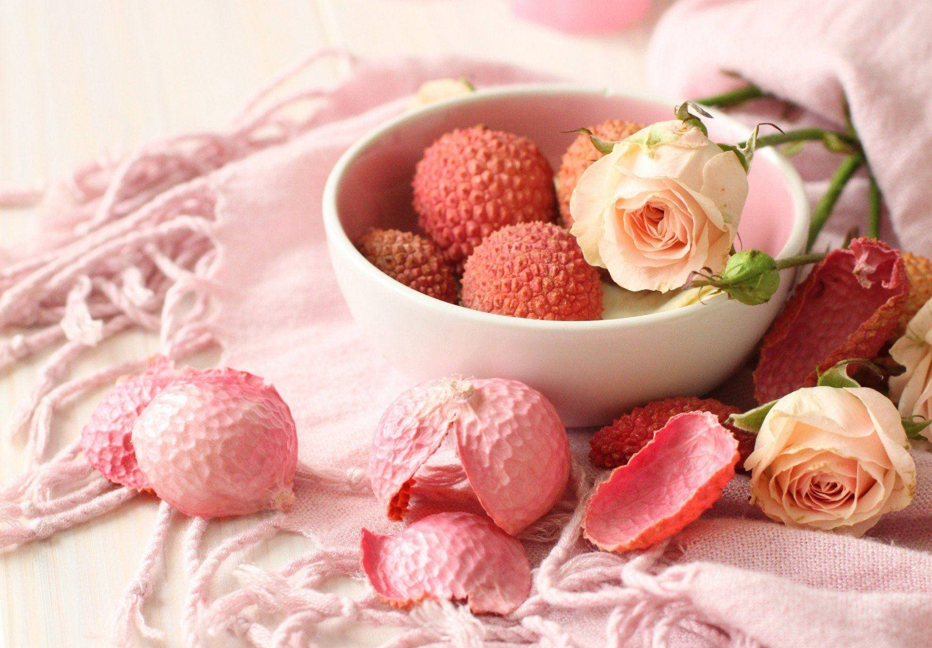 Litchi 桌布 壁紙 — 一個白色碗盛裝新鮮荔枝和淡粉紅玫瑰，放在柔軟的粉紅色流蘇布上，周圍散落著荔枝皮。