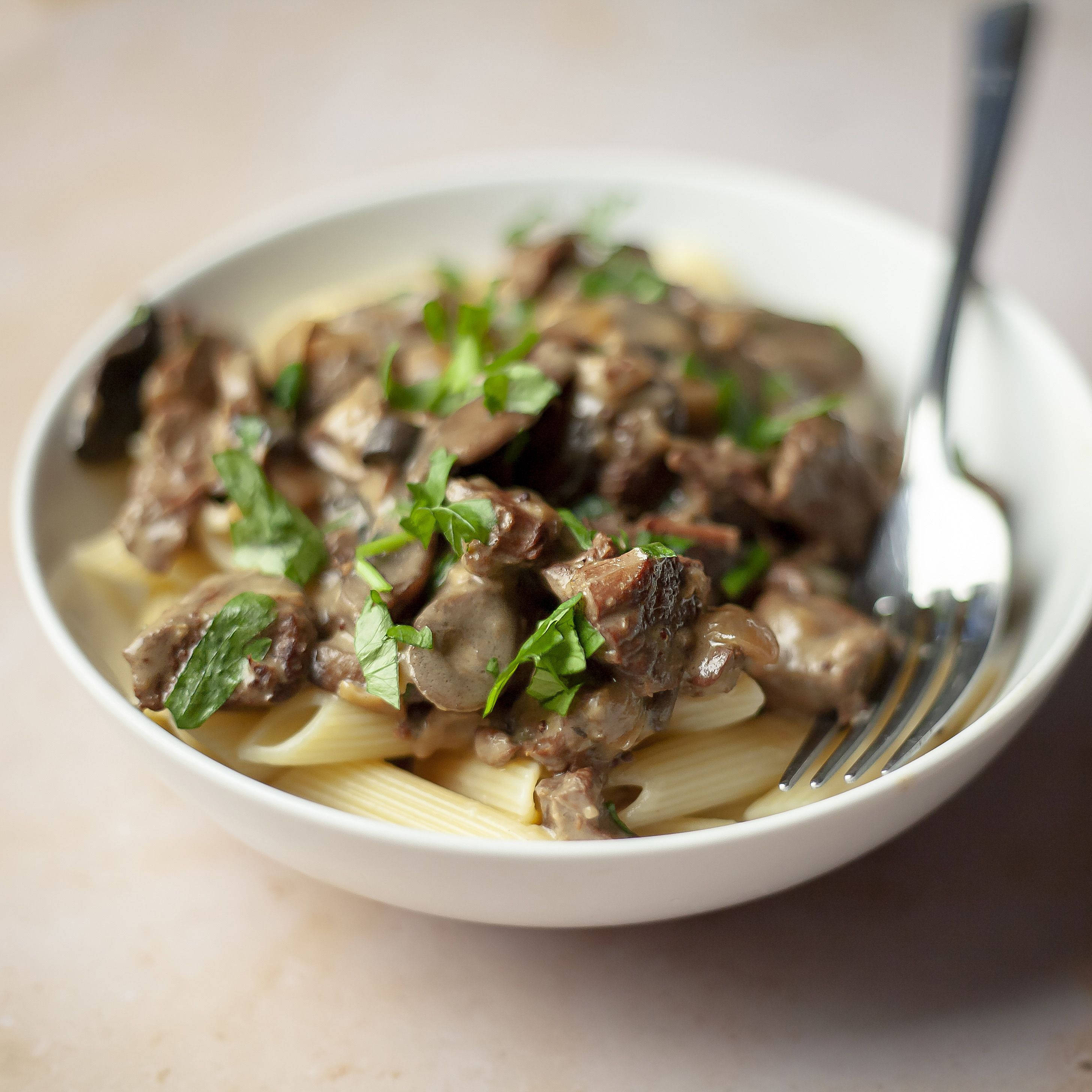 Food Photography 桌布 壁紙 — 一個白色碗盛裝著奶油牛肉斯特羅根諾夫，搭配蘑菇和新鮮歐芹，放在筆管麵上，旁邊擱著叉子。