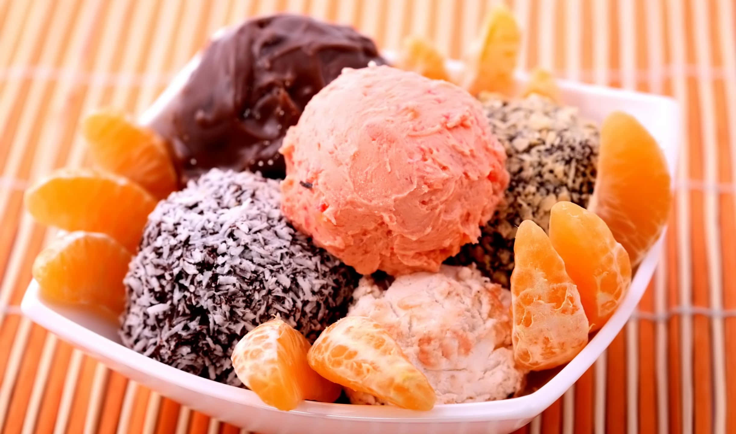 2440x1440 Desserts 桌布 壁紙 — 一個白色碗盛載著各種色彩繽紛的冰淇淋球和柑橘橙片，放在竹蓆上。