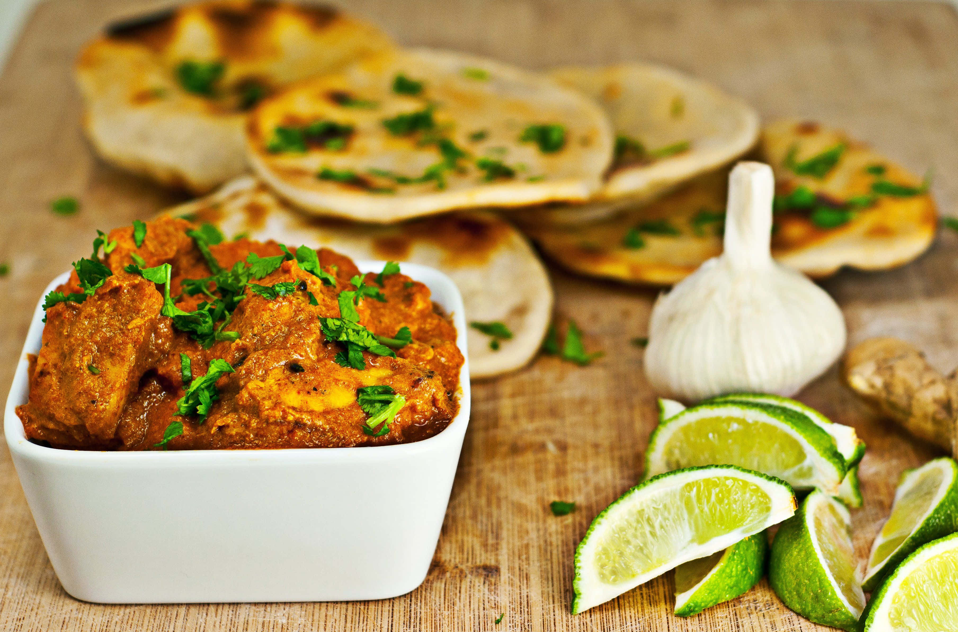 Indian Food 桌布 壁紙 — 一個白色碗盛載著濃郁的橙色咖哩，配上香菜裝飾，旁邊有萊姆角、大蒜和印度烤餅放在木板上。