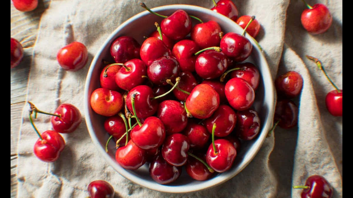 Red Cherries 桌布 壁紙 — 一個白色碗裝滿了光澤的紅色櫻桃，放在有紋理的米色布上，周圍散落著櫻桃。