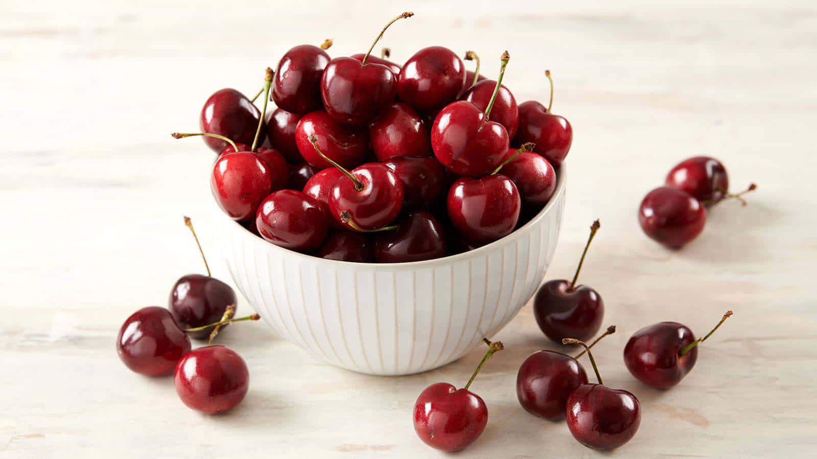 Red Cherries 桌布 壁紙 — 一個白色碗裝滿了光澤的紅色櫻桃，放在淺色木質表面上，周圍散落著水果。