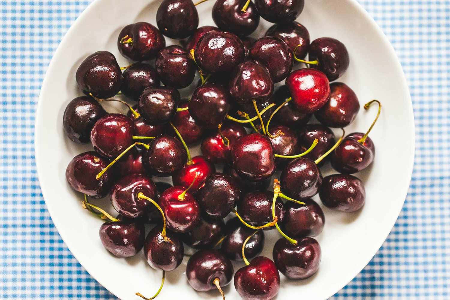 Red Cherries 桌布 壁紙 — 一個白色碗裝滿了帶有綠色莖的光澤深紅色櫻桃，放在藍白格子桌布上。