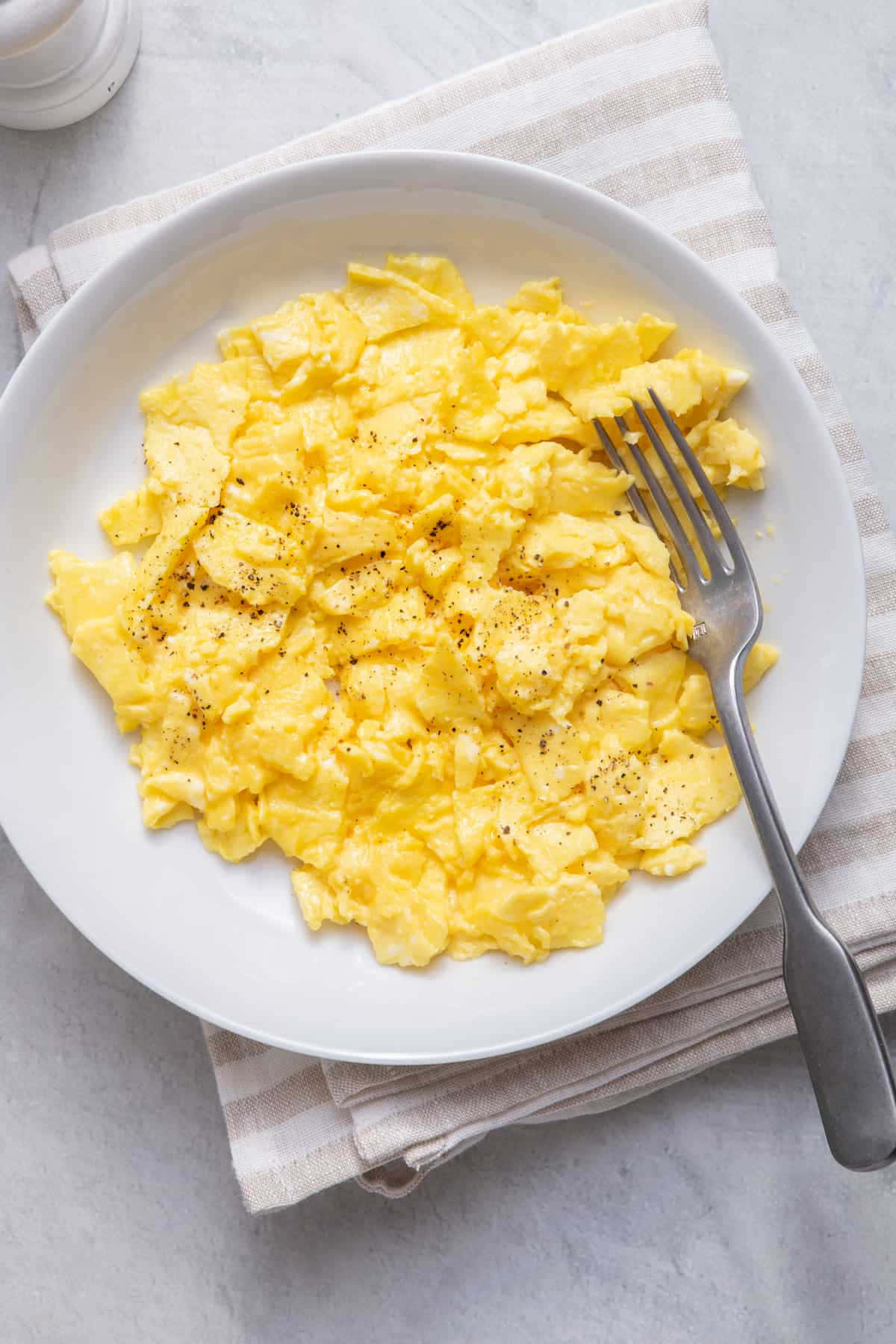 Scrambled Egg 桌布 壁紙 — 一個白色碗裝滿了鬆軟的黃色炒蛋，撒上黑胡椒，放在條紋餐巾上，旁邊有一把叉子。