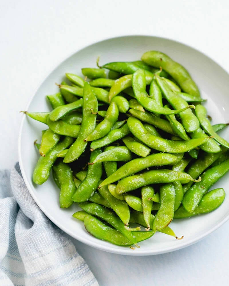 Vegetables 桌布 壁紙 — 一個白色碗裝滿了鮮綠色鹽味毛豆莢，放在條紋布上，背景是乾淨的白色。
