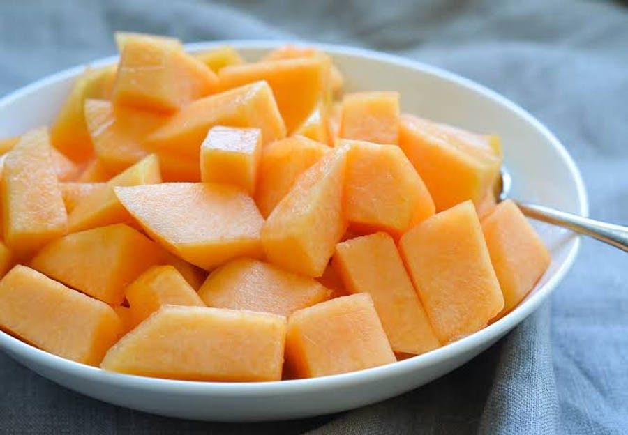 Fruit 桌布 壁紙 — 一個白色碗裝滿多汁的橙色哈密瓜塊，放在灰色布上，旁邊放著一隻湯匙。