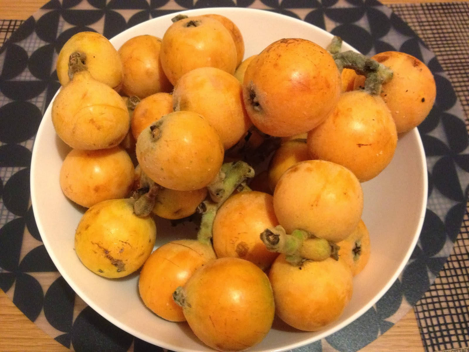 Fruits 桌布 壁紙 — 一個白色碗裝滿成熟金黃色枇杷，放在有圖案的桌布上，在溫暖的室內燈光下。