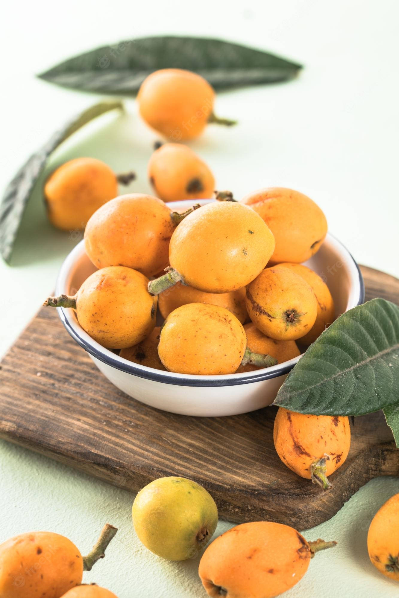 Fruits 桌布 壁紙 — 一個白色碗裝滿成熟金黃色枇杷，放在一塊鄉村風格木板上，周圍有新鮮綠葉和散落的水果。