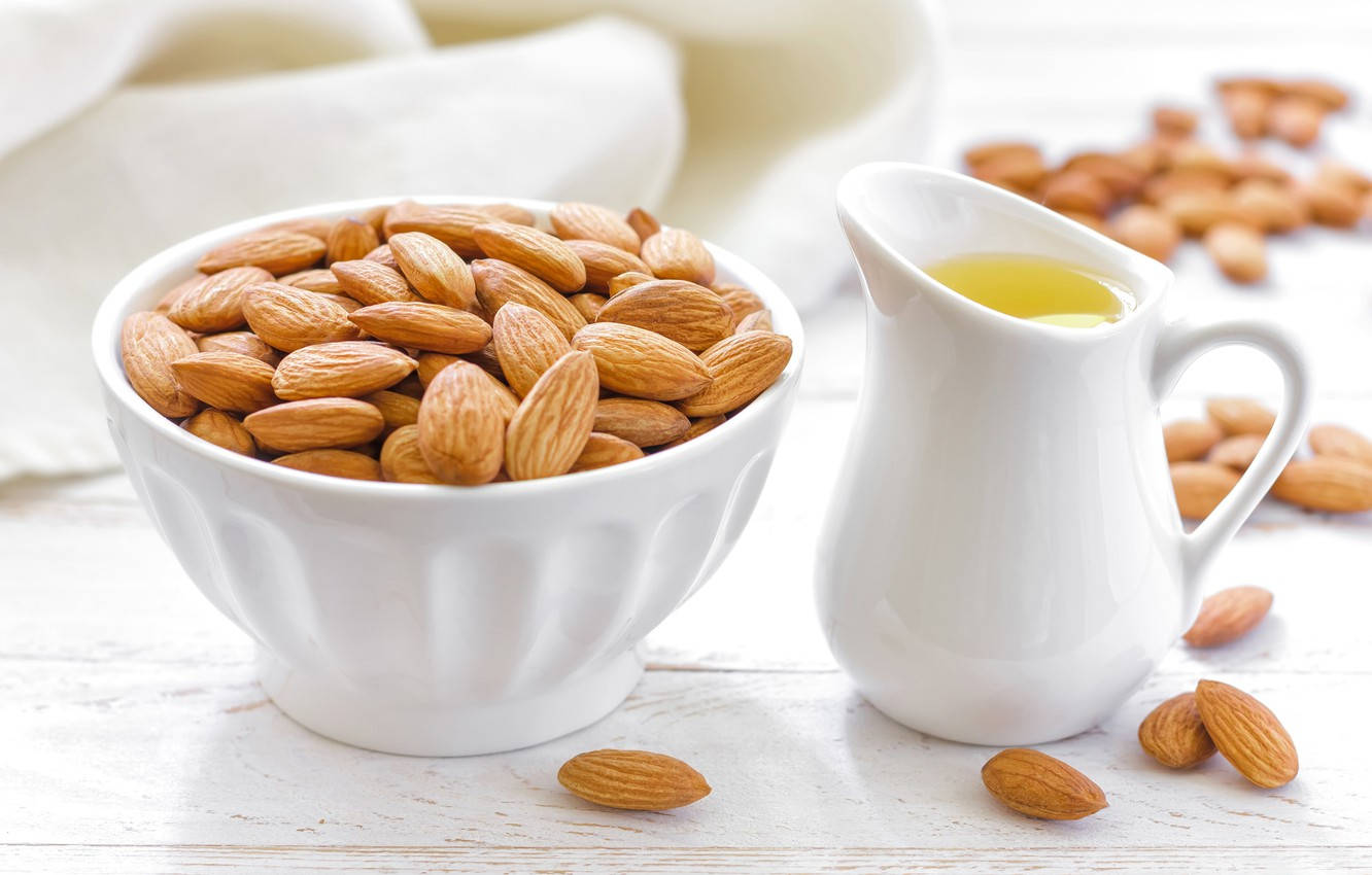Almond 桌布 壁紙 — 一個白色碗裝滿杏仁，旁邊放著一壺杏仁油，放在質樸的木桌上。