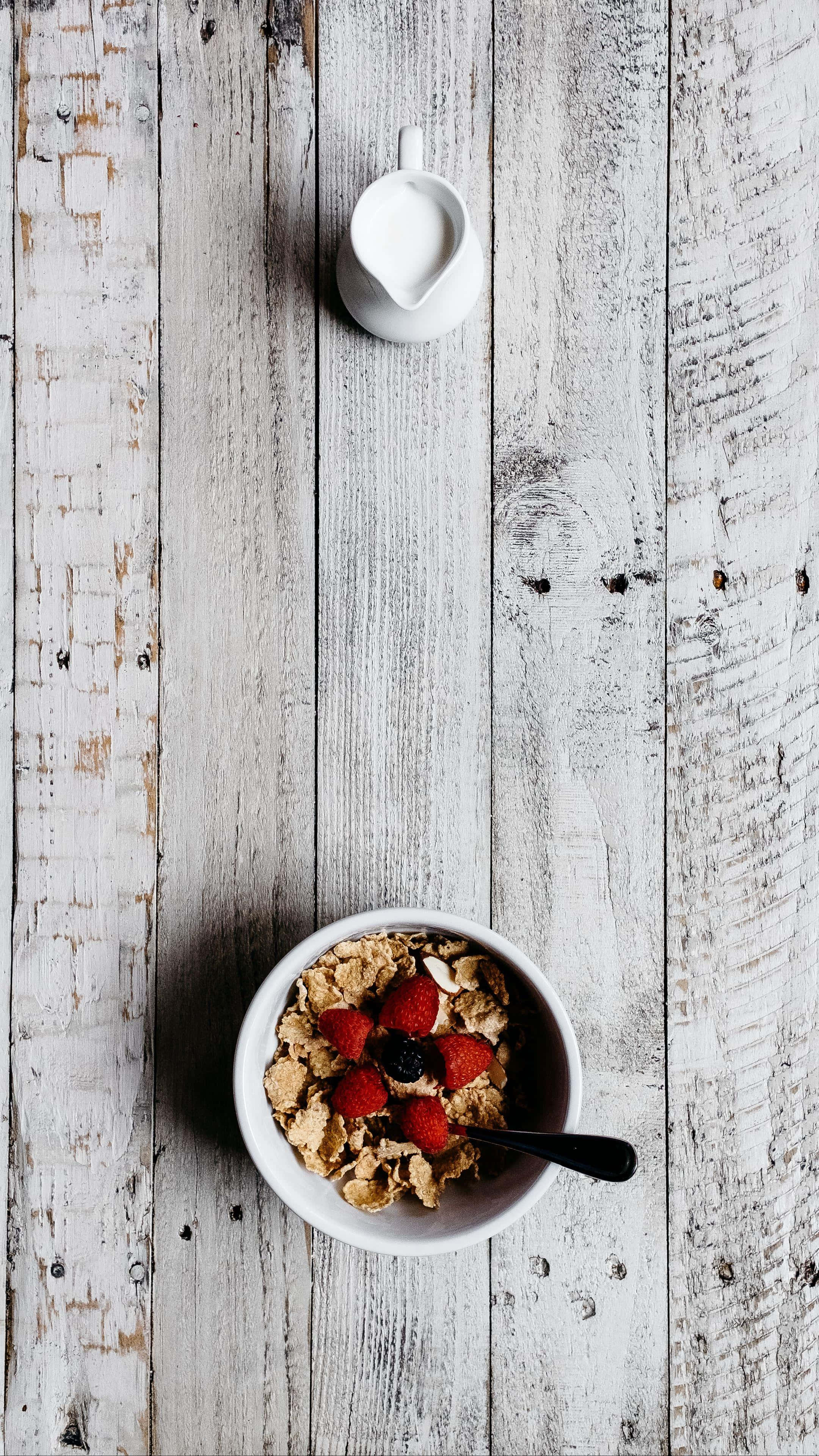 Cereal Bowl 桌布 壁紙 — 一個白色碗裝滿穀物和新鮮草莓，旁邊有一個小牛奶壺，放在一個風化的白色木桌上，從上方俯瞰的視角。