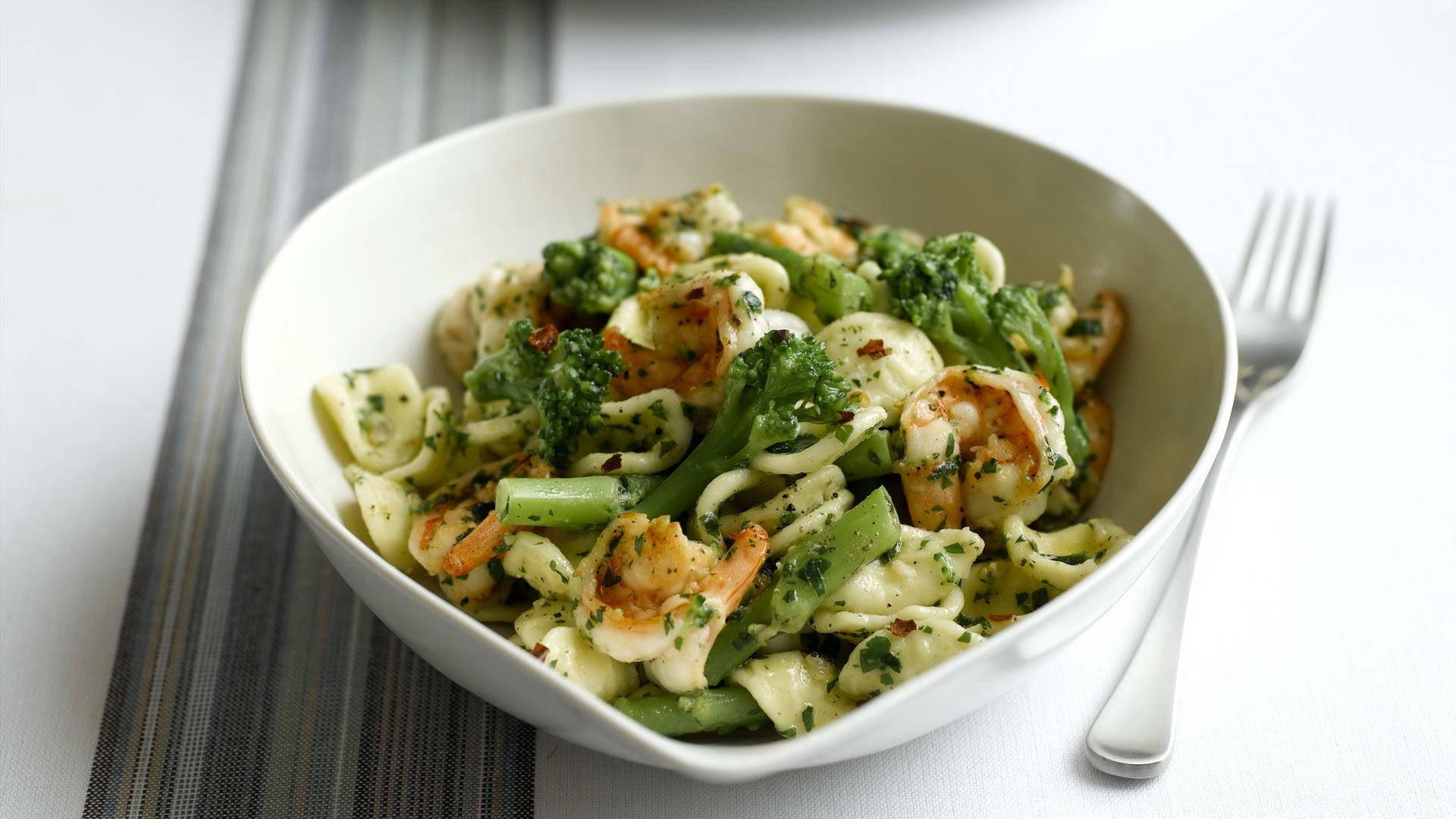 Pasta 桌布 壁紙 — 一個白色碗裝滿義大利麵、蝦子和青花菜，拌上綠色香草醬，放在條紋桌布上，旁邊有叉子。