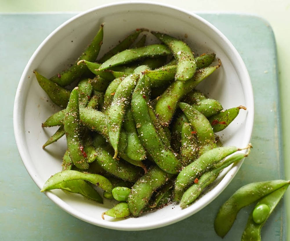 beans 桌布 壁紙 — 一個白色碗裝滿調味綠毛豆莢，放在淡綠色表面上，附近散落幾個豆莢。