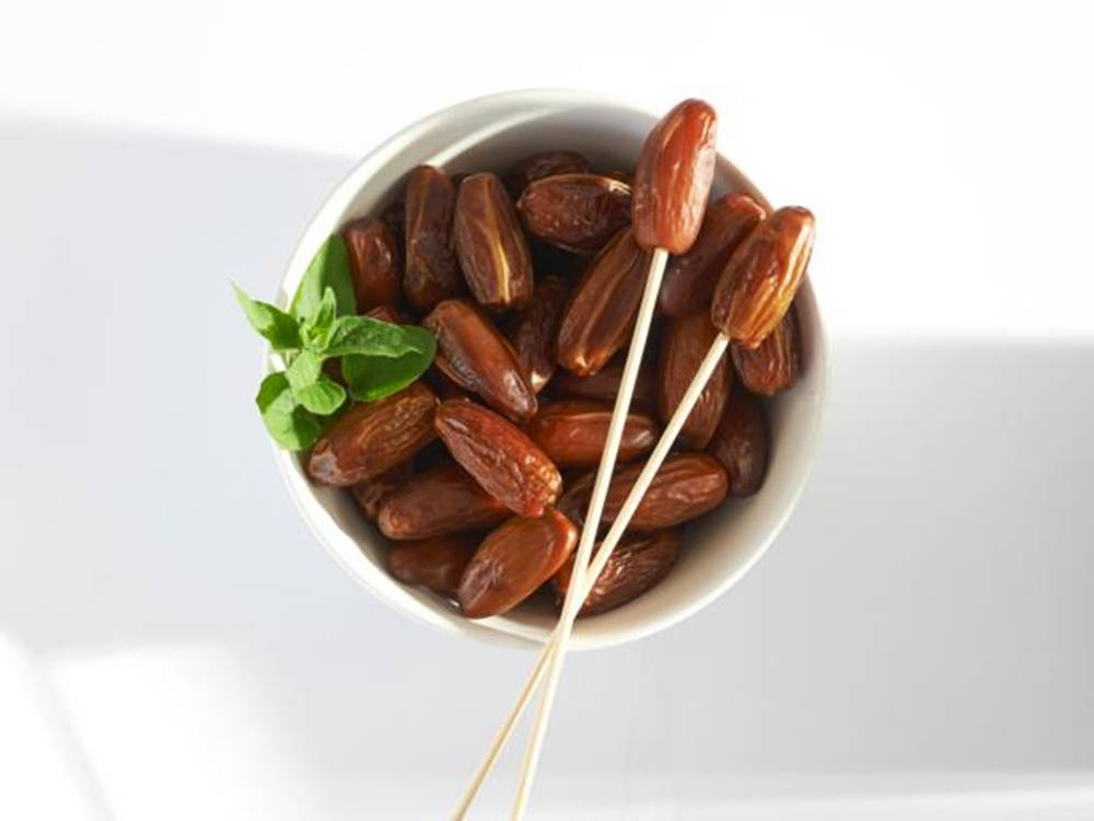 美學 桌布 壁紙 — 一個白色碗裝滿飽滿的棕色椰棗，飾以新鮮綠色薄荷葉和兩根竹籤。