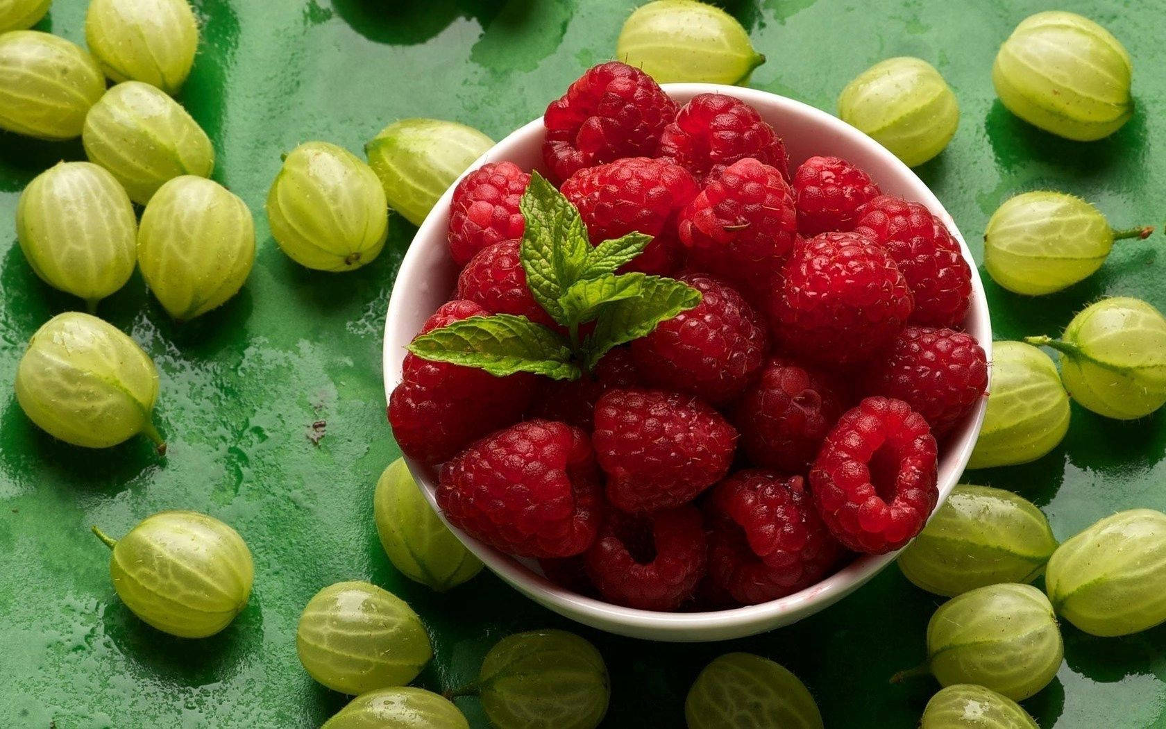 Fruit 桌布 壁紙 — 一個白色碗裝滿鮮紅色的覆盆子和新鮮薄荷，放在濕潤的綠色表面上，周圍環繞著淡綠色的醋栗。