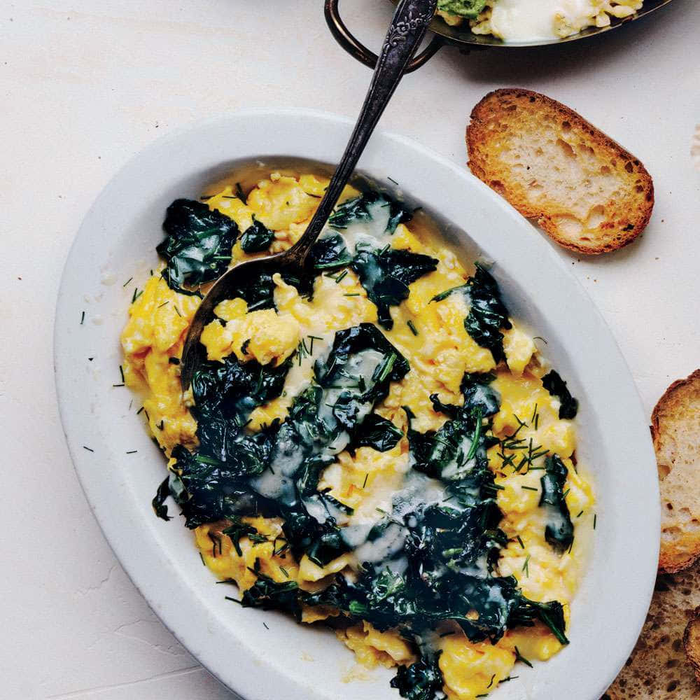 Scrambled Egg 桌布 壁紙 — 一個白色碗裝著奶油狀炒蛋，混合了萎蔫的菠菜和蒔蘿，旁邊是放在淺色表面上的烤麵包。