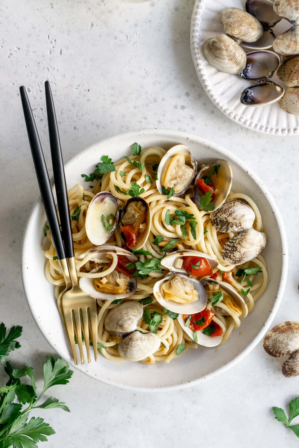 Clam 桌布 壁紙 — 一個白色碗裝蛤蜊義大利麵，搭配櫻桃番茄和歐芹，放在有斑點的檯面上，搭配黑色和金色餐具。