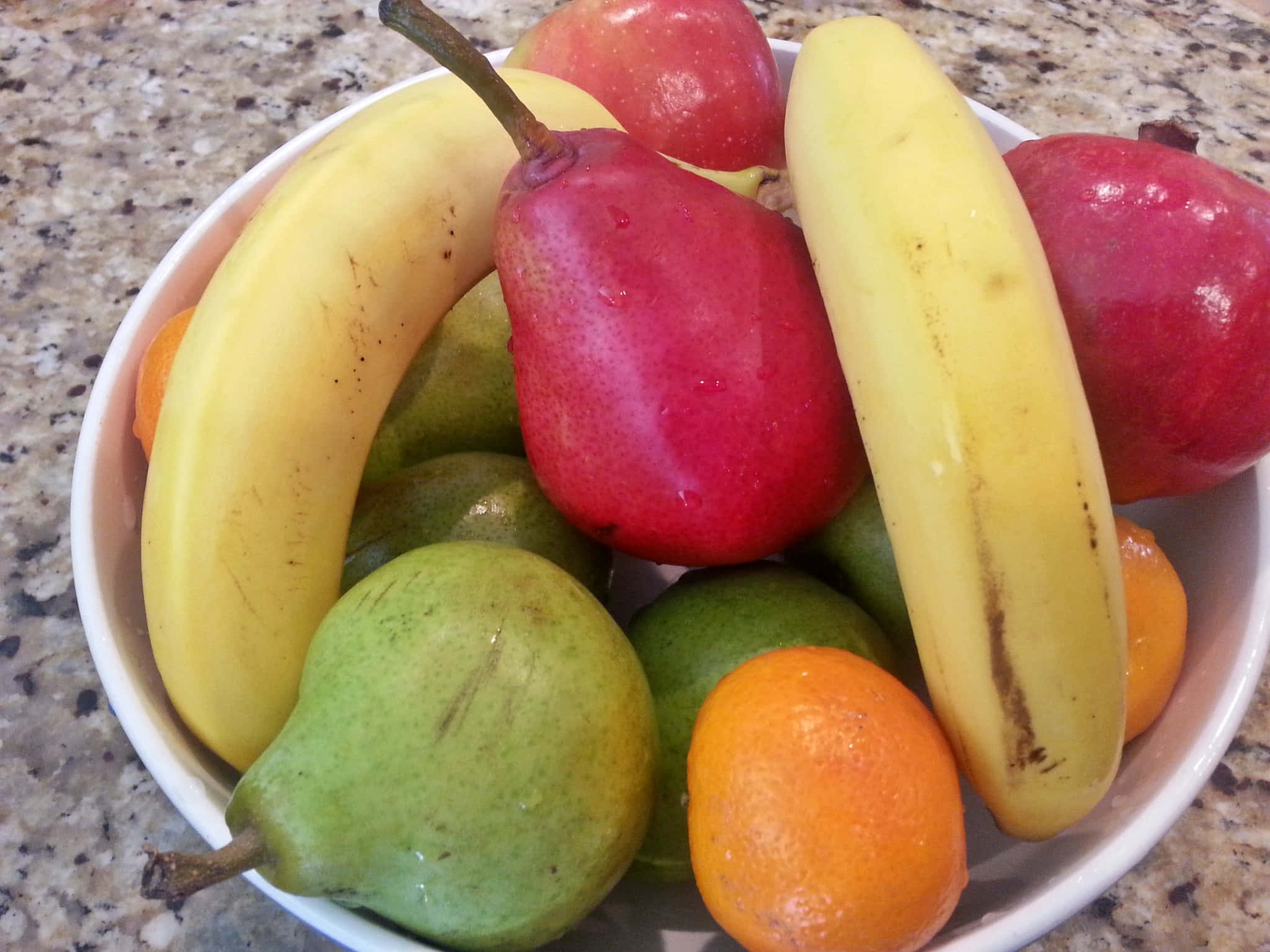 Winter Fruits 桌布 壁紙 — 一個白色碗裡裝滿了鮮黃色的香蕉、紅色的蘋果、綠色的梨和橙色的柑橘類水果，放在一個有斑點的檯面上。