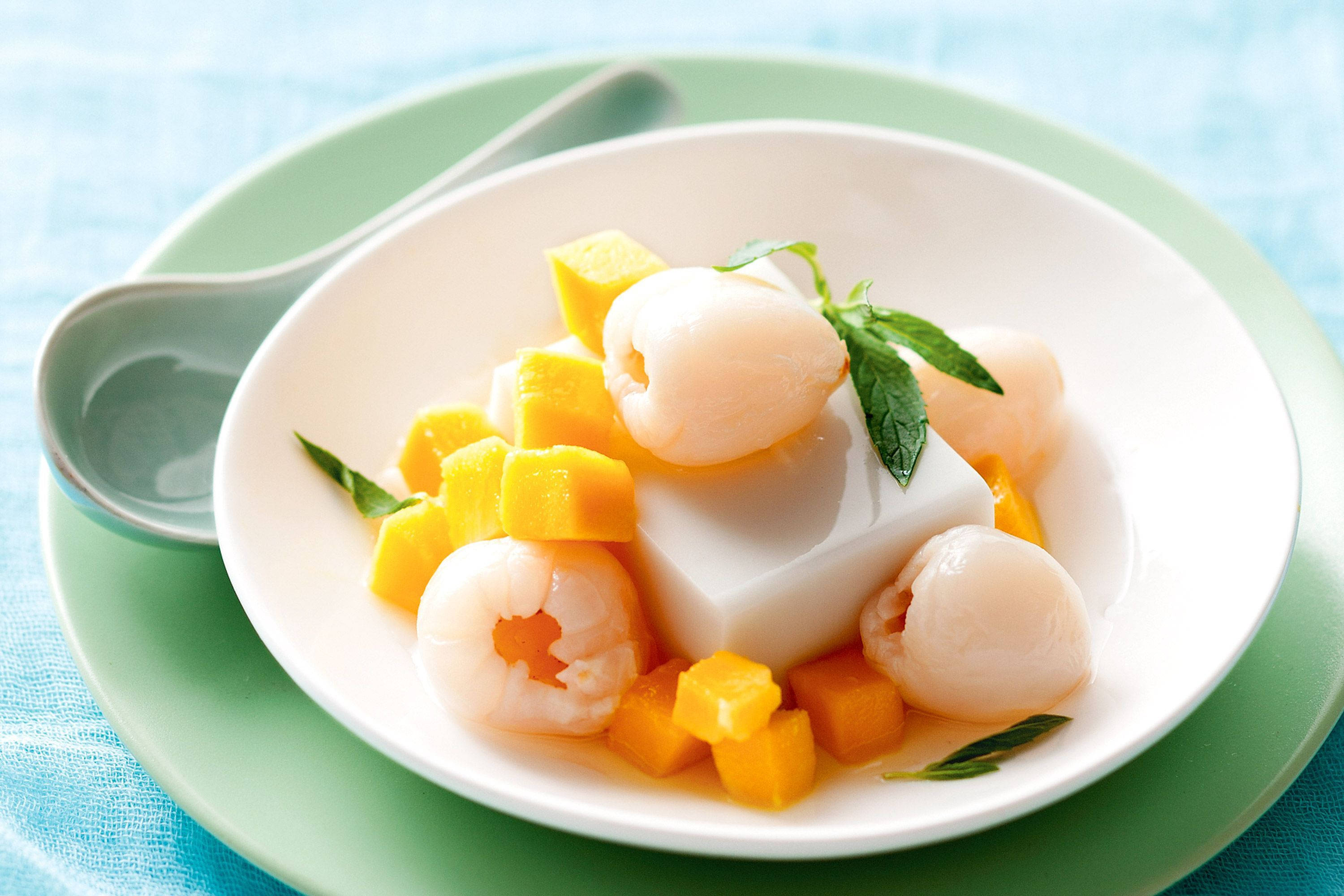 Litchi 桌布 壁紙 — 一個白色碗裡裝著半透明的甜椰子果凍方塊，上面放著多汁的黃色芒果塊和飽滿的荔枝，並用薄荷葉裝飾。