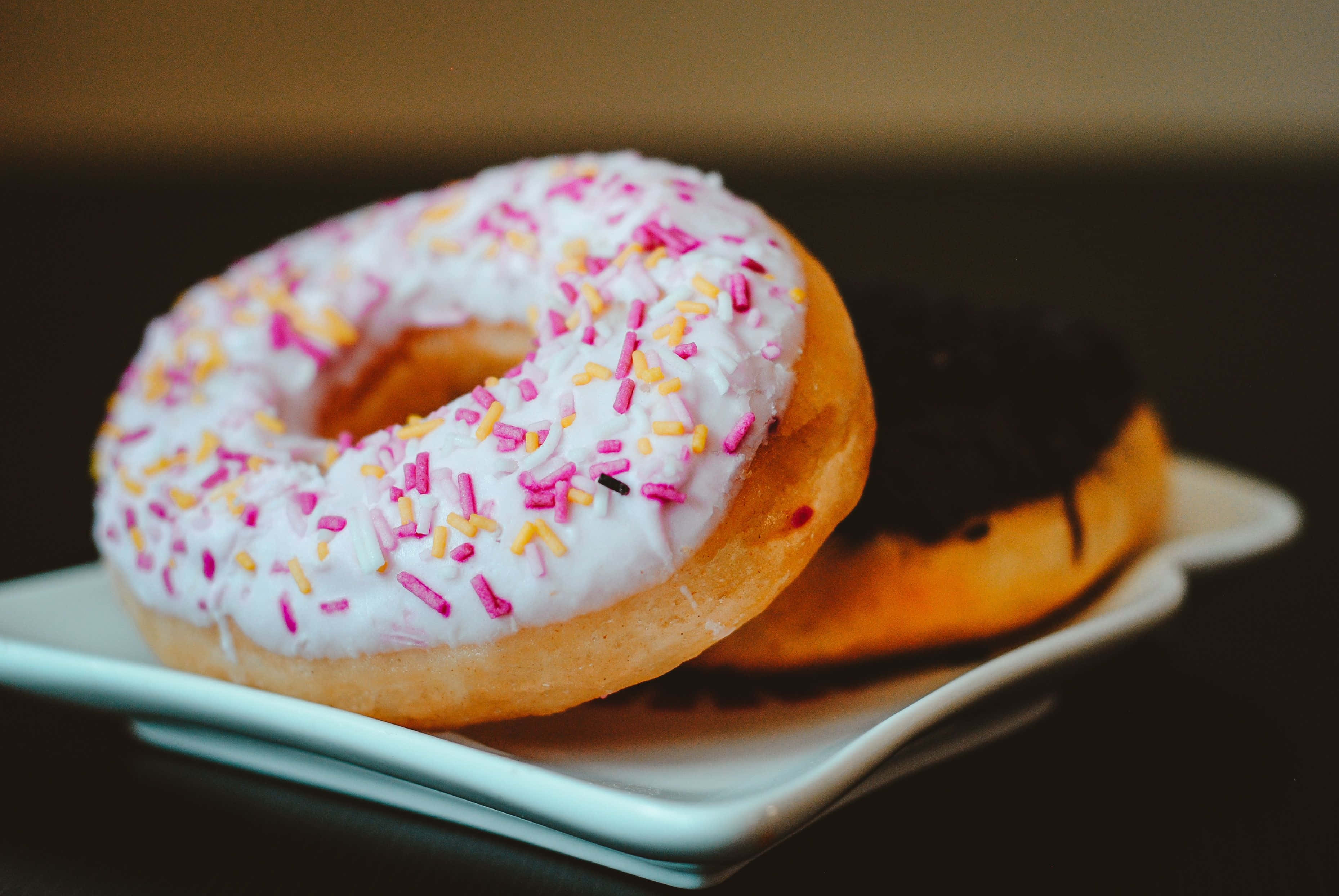 Dunkin Donuts 桌布 壁紙 — 一個白色糖霜甜甜圈配上粉紅色和黃色糖針，旁邊有一個巧克力甜甜圈，放在白色盤子上的特寫。