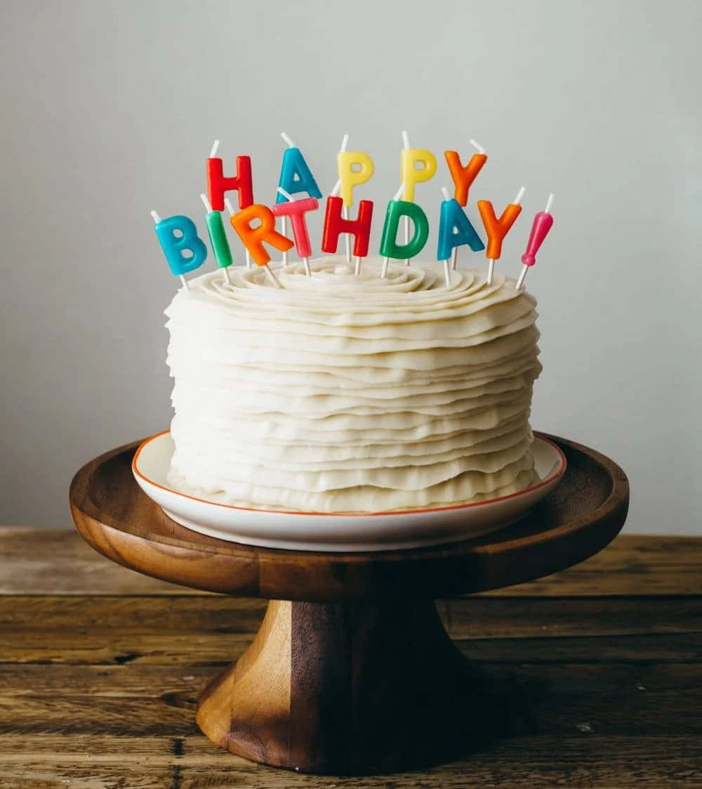 Happy Birthday Cake 桌布 壁紙 — 一個白色糖霜生日蛋糕，上面有彩色字母蠟燭拼出 happy birthday，放在木製蛋糕架上。