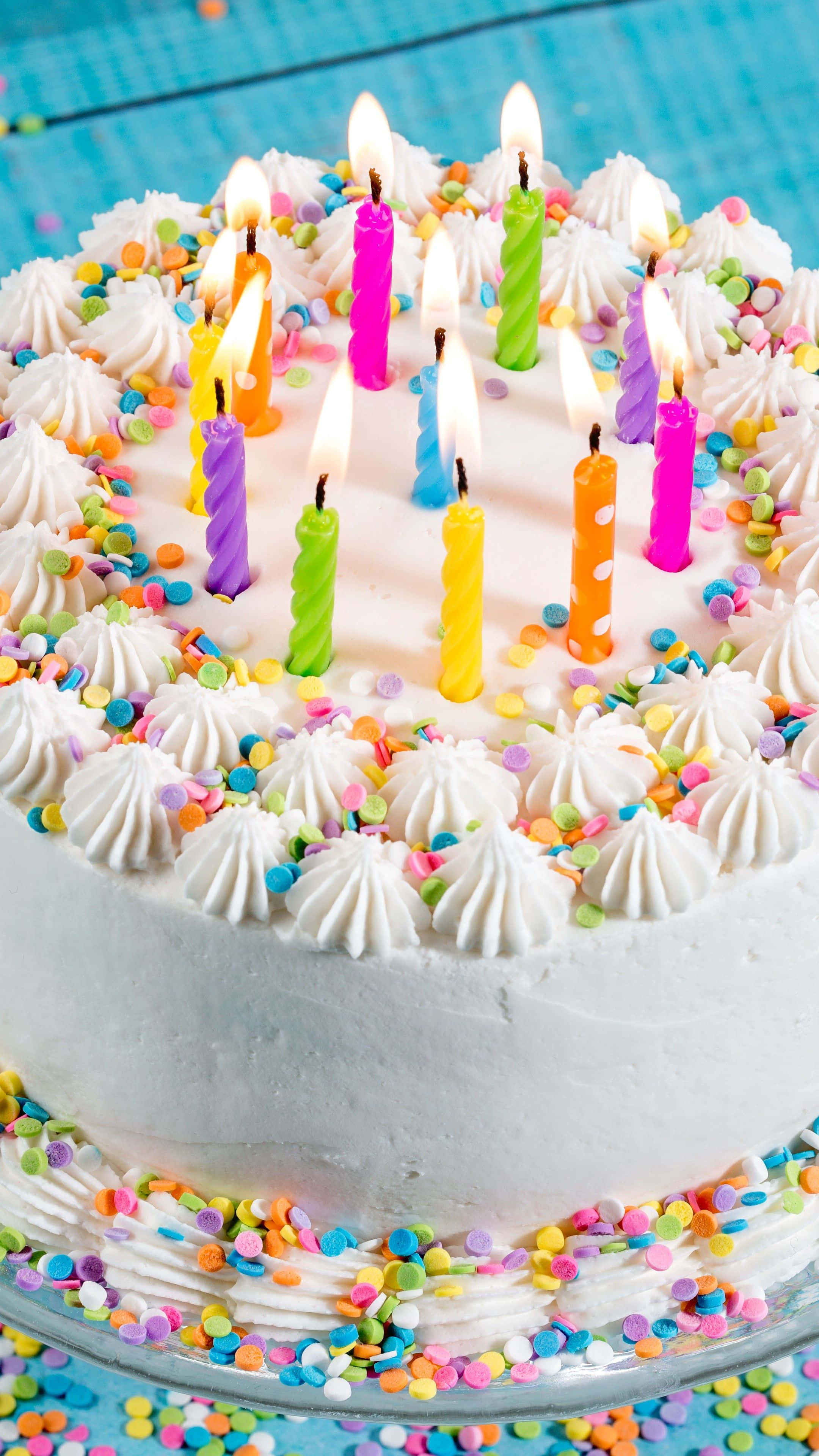 Birthday Cakes 桌布 壁紙 — 一個白色糖霜生日蛋糕的特寫，上面裝飾著彩色蠟燭和彩虹糖針，背景是藍色格子圖案。