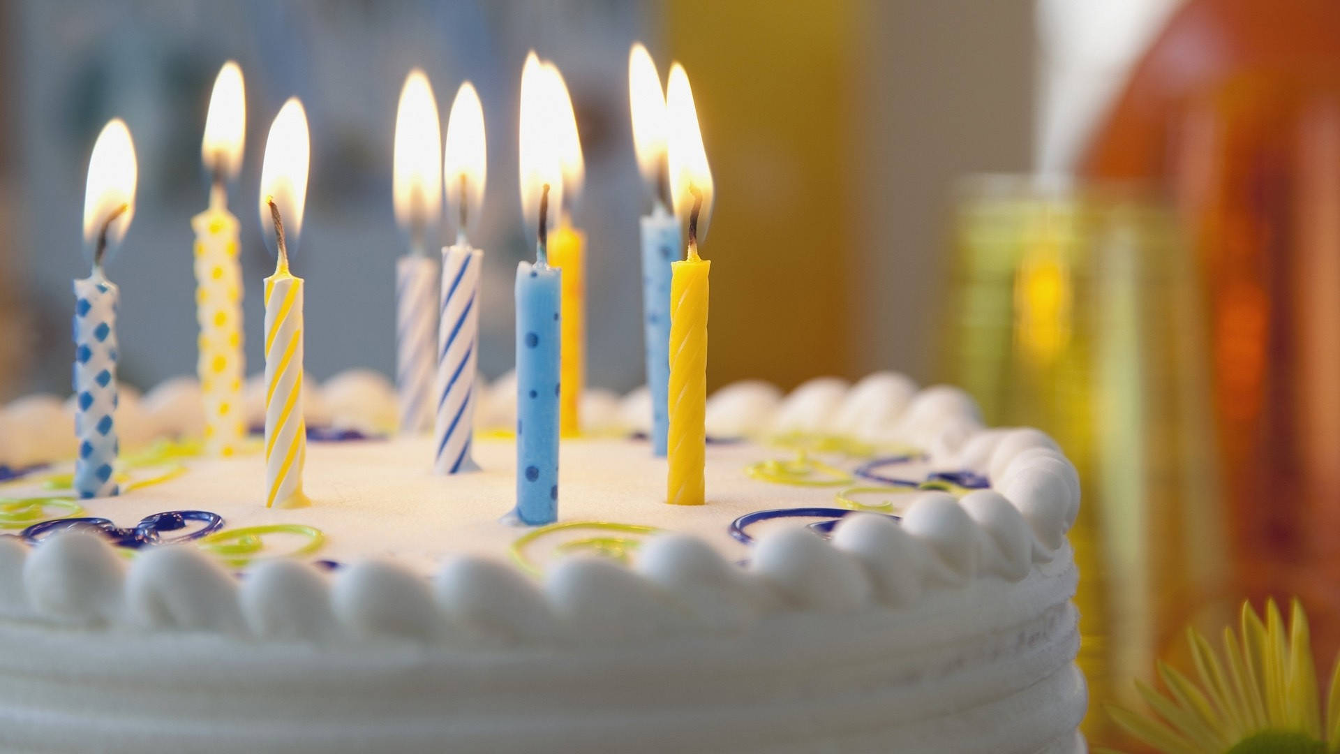 Cake 桌布 壁紙 — 一個白色糖霜生日蛋糕的特寫，裝飾著點燃的藍色、黃色和條紋蠟燭，散發出溫暖發光的火焰。