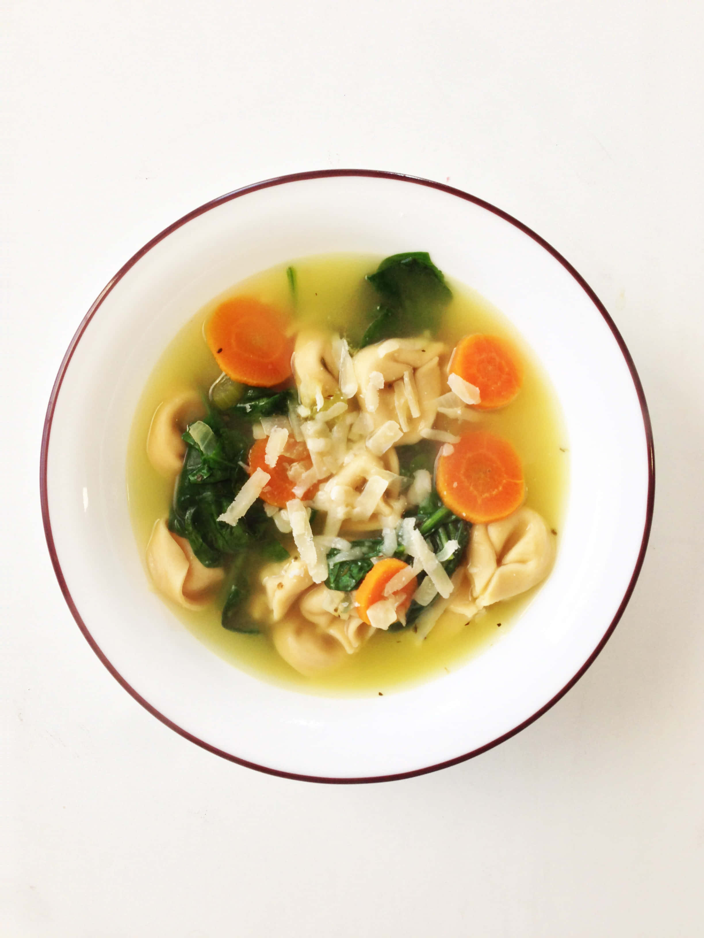Pasta 桌布 壁紙 — 一個白色紅邊碗裝滿了義大利餃子湯，上面放有菠菜、胡蘿蔔片和磨碎起司的俯視圖。